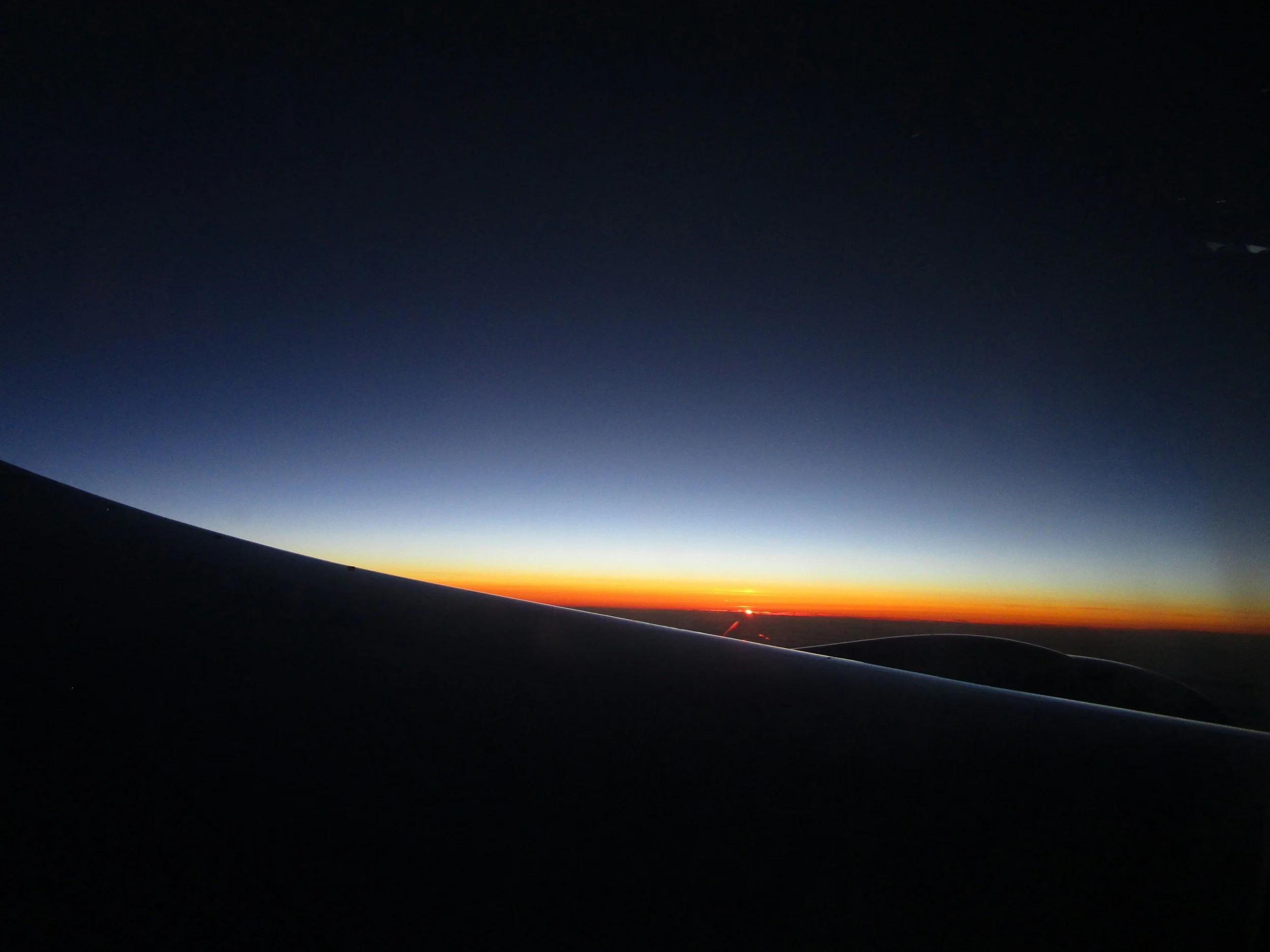 Vista de una puesta de sol o un amanecer desde la ventanilla de un avión, en la que se ve el ala del avión en primer plano y el horizonte, con intensos tonos naranjas, rojos y amarillos que se funden con el cielo azul y negro.