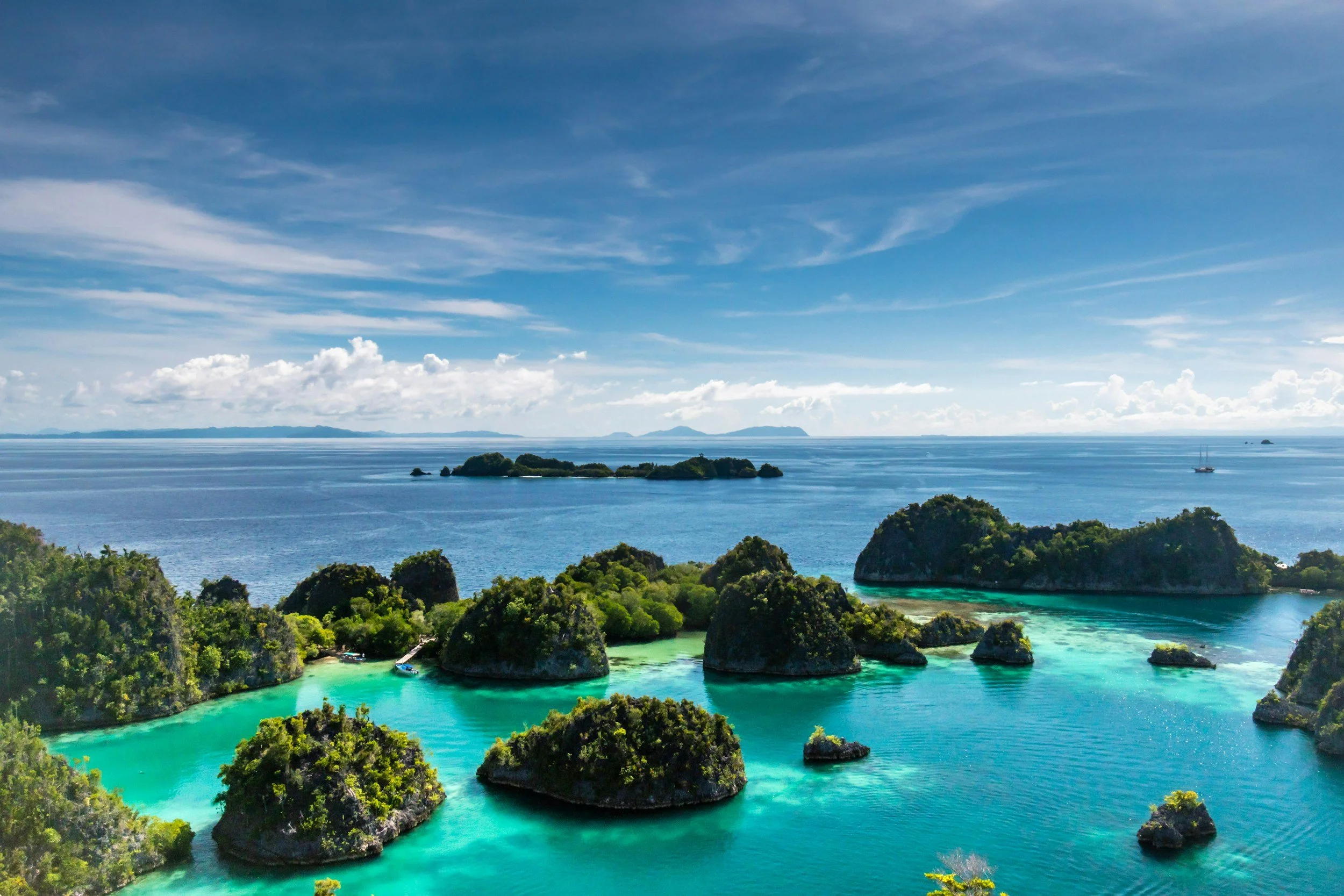 Una vista panorámica de aguas turquesas con pequeñas islas y frondosos árboles verdes en primer plano, una gran extensión de agua con un velero en la distancia y un cielo parcialmente nublado en lo alto.