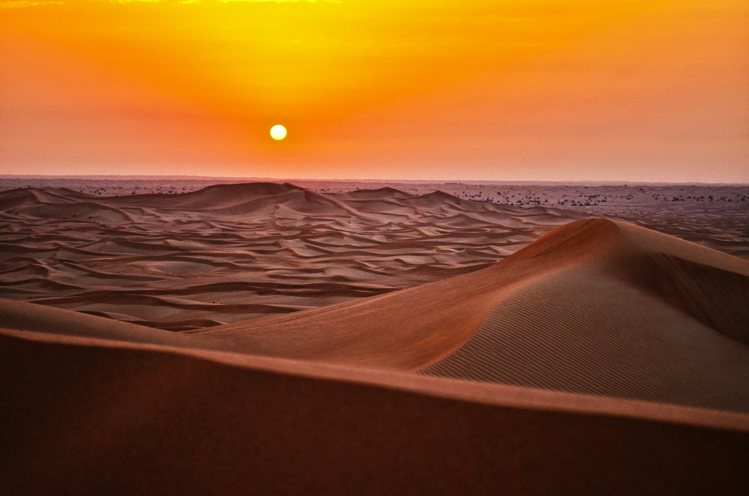 Un paisaje desértico al atardecer, con dunas en primer plano y el sol poniéndose en el horizonte, creando un cielo cálido en tonos naranjas y amarillos.
