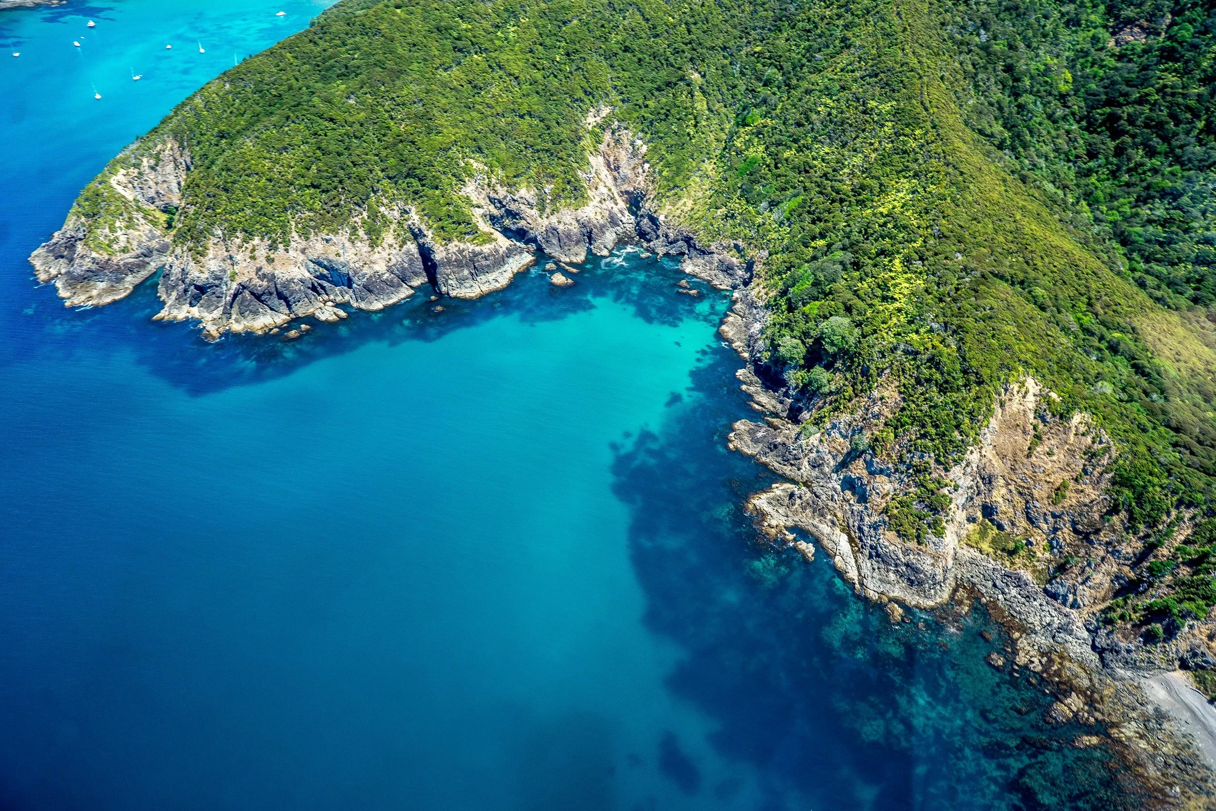 Vista aérea de una isla frondosa y verde rodeada por las aguas azul brillante del océano, con pequeños veleros en la distancia.