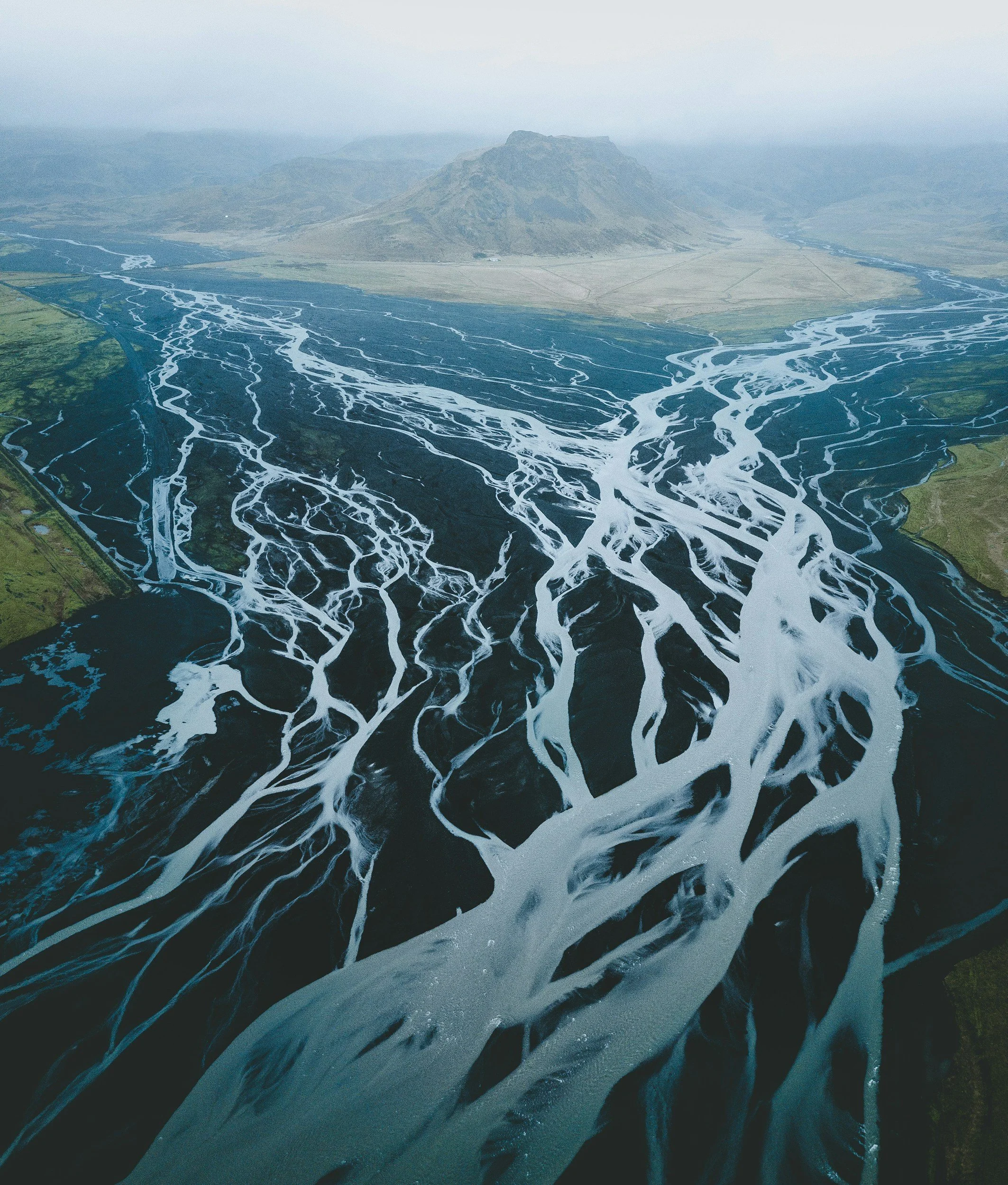 Vista aérea de un delta fluvial con múltiples canales de agua que atraviesan una zona verde y desembocan en una montaña al fondo, bajo un cielo nublado.