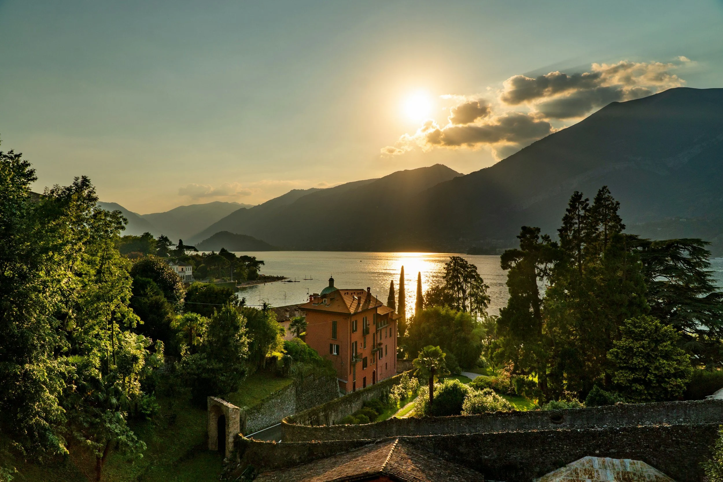 Una vista panorámica de una puesta de sol sobre un lago rodeado de montañas y frondosos árboles, con un edificio rosa y antiguas construcciones de piedra en primer plano.