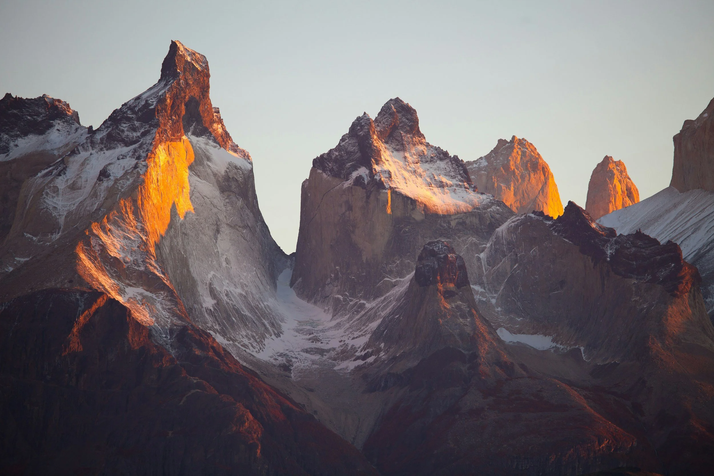 Montañas nevadas con picos rocosos iluminados por el sol poniente o naciente, con un cielo despejado de fondo.
