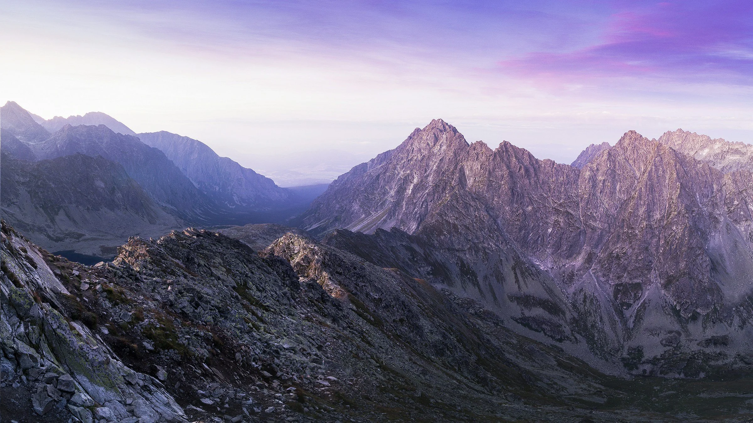 Una vista panorámica de escarpadas cumbres montañosas al atardecer, con un cielo teñido de tonos morados y rosados.
