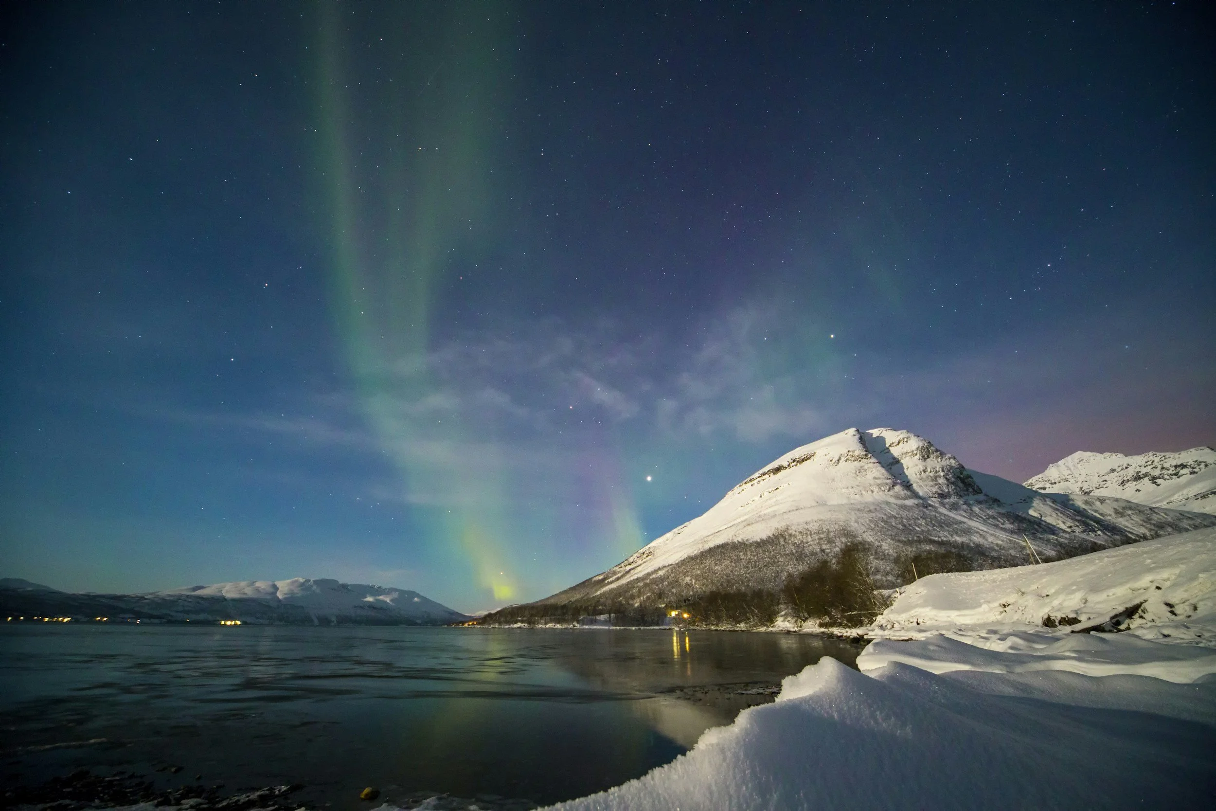 Nighttime scene of a snow-covered mountain range with the northern lights (aurora borealis) in green hues across the dark sky, reflections in the water, and snow at the shore.