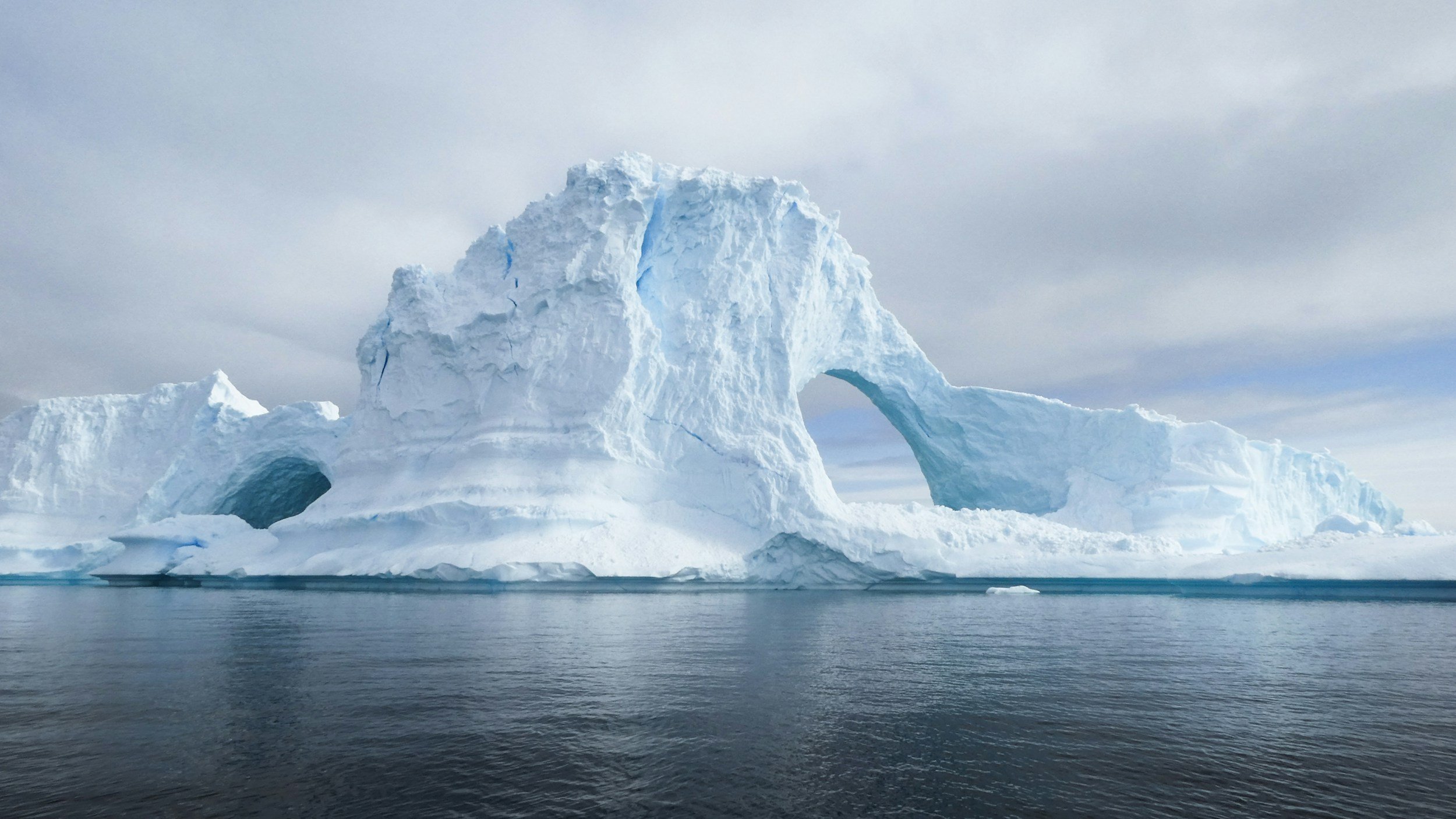 A large iceberg with a natural arch, floating in cold ocean water under a cloudy sky.