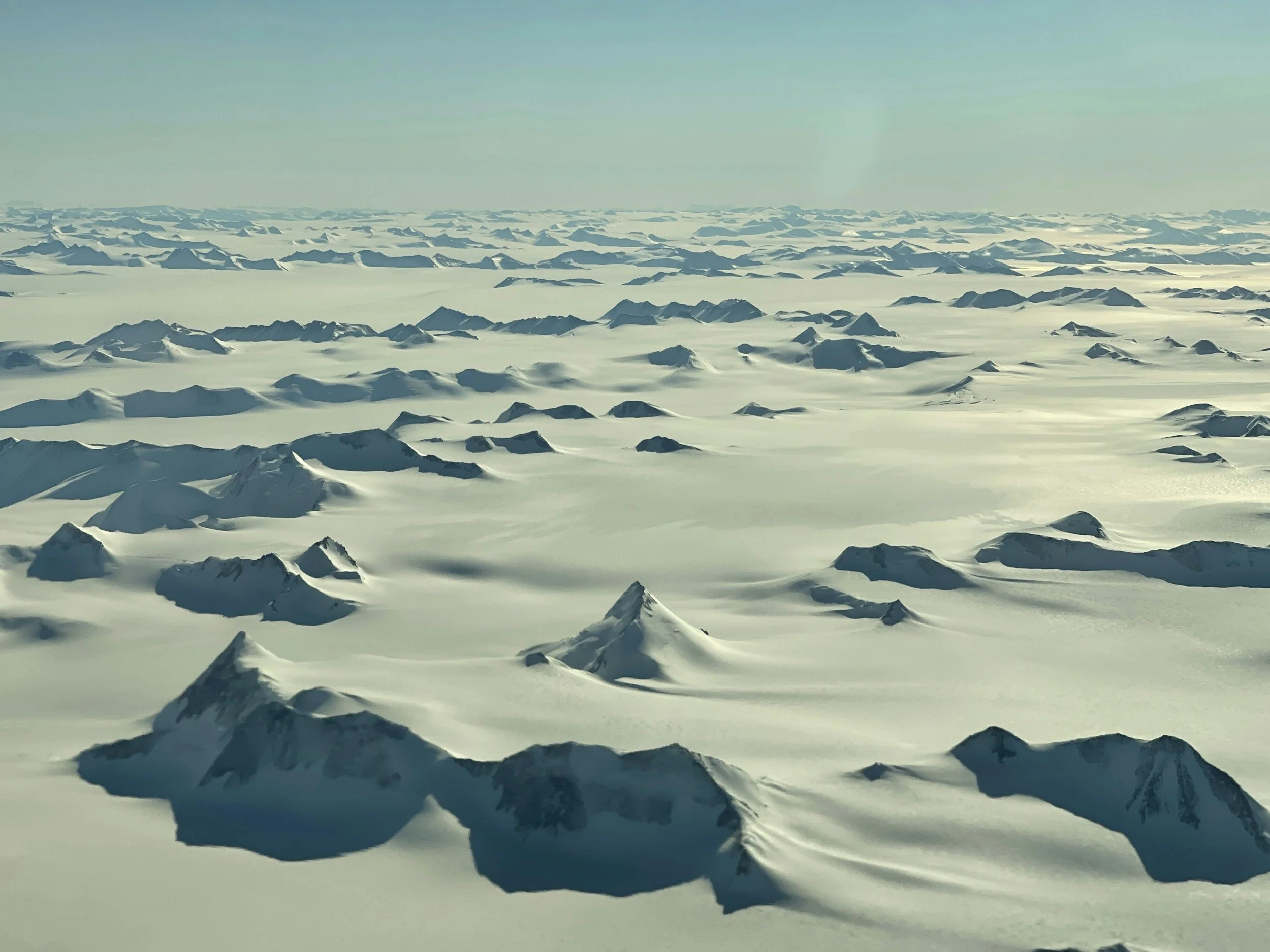 Snow-covered mountain range extending to the horizon under a pale blue sky.