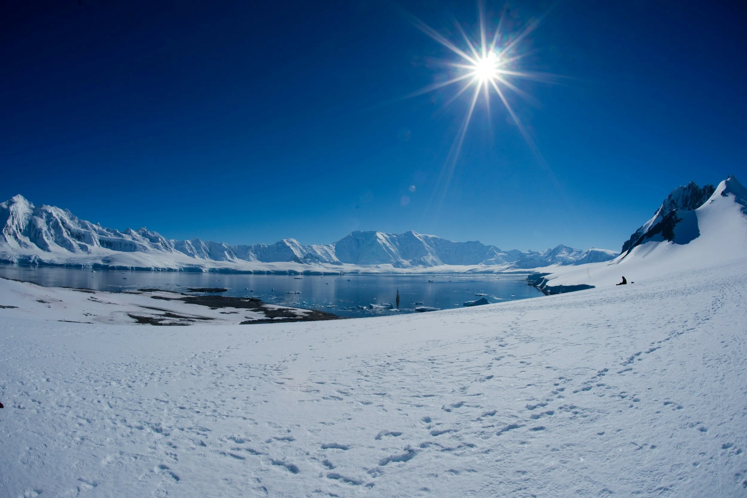 Snow-covered landscape with a mountain range in the distance, a partially frozen body of water, bright sunlight in a clear blue sky, and a small figure sitting on the snow in the foreground.