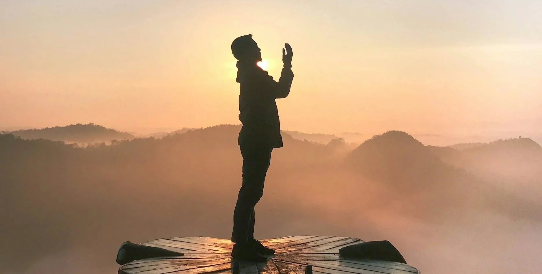 Silhouette of a person standing on a wooden platform with hands together in a prayer position, against a backdrop of a misty mountain landscape during sunrise or sunset.