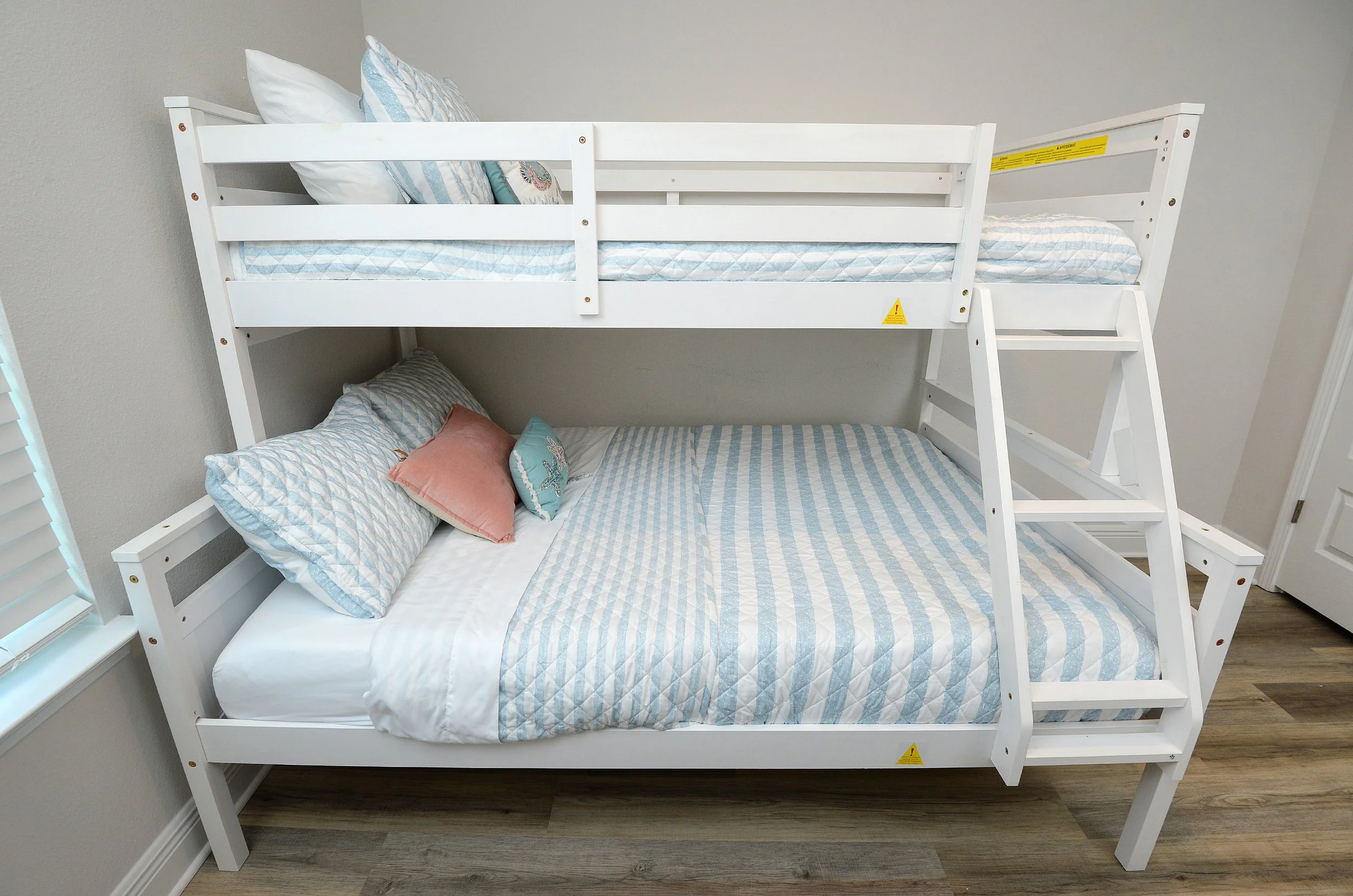 White bunk bed with blue and white striped bedding and multiple pillows in a bedroom with gray walls and wood flooring.