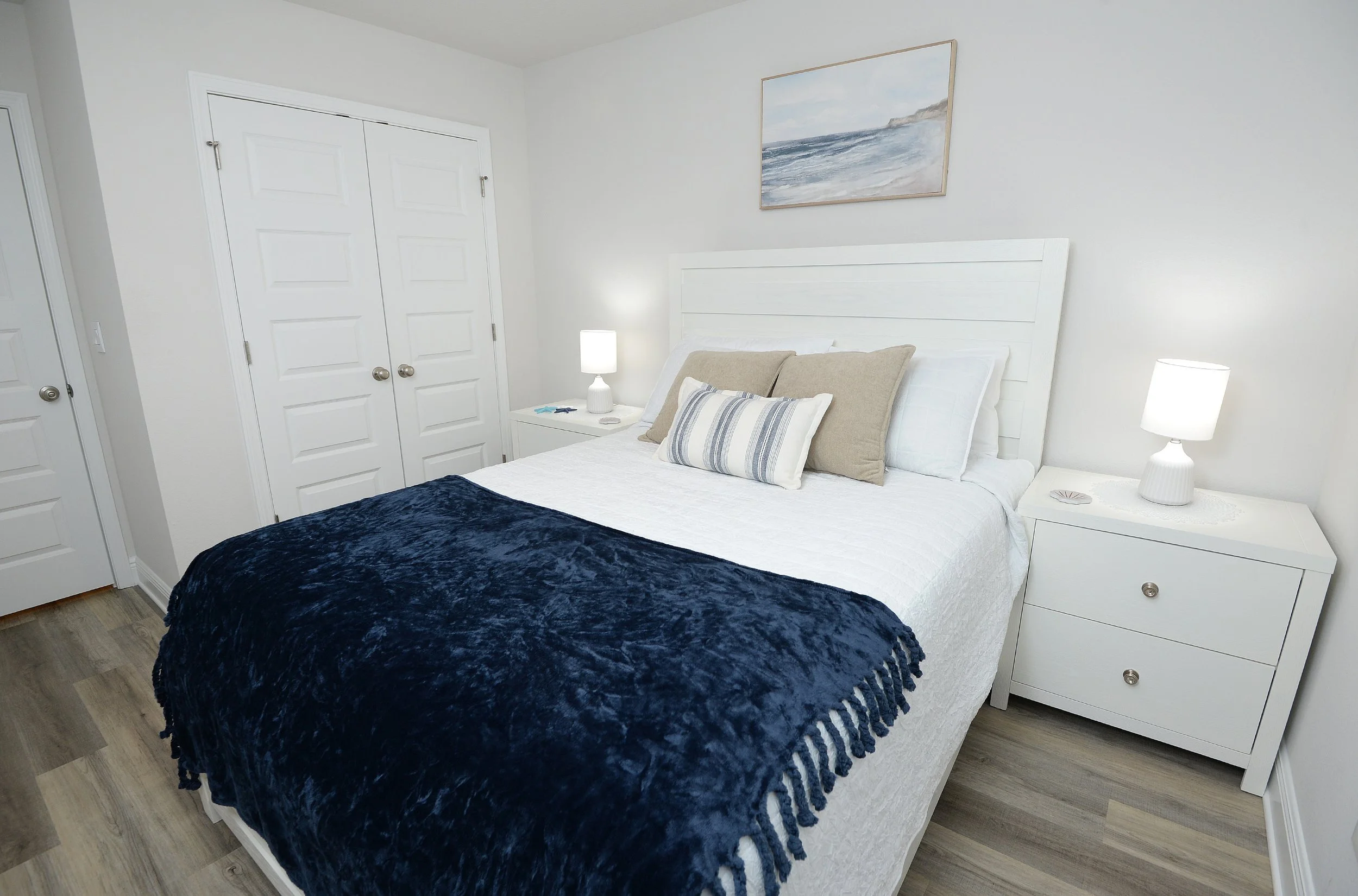 A bedroom with white walls, a white bed with beige and white pillows, a navy blue blanket, white nightstands with lamps, a painting of the ocean on the wall, and a wooden floor.