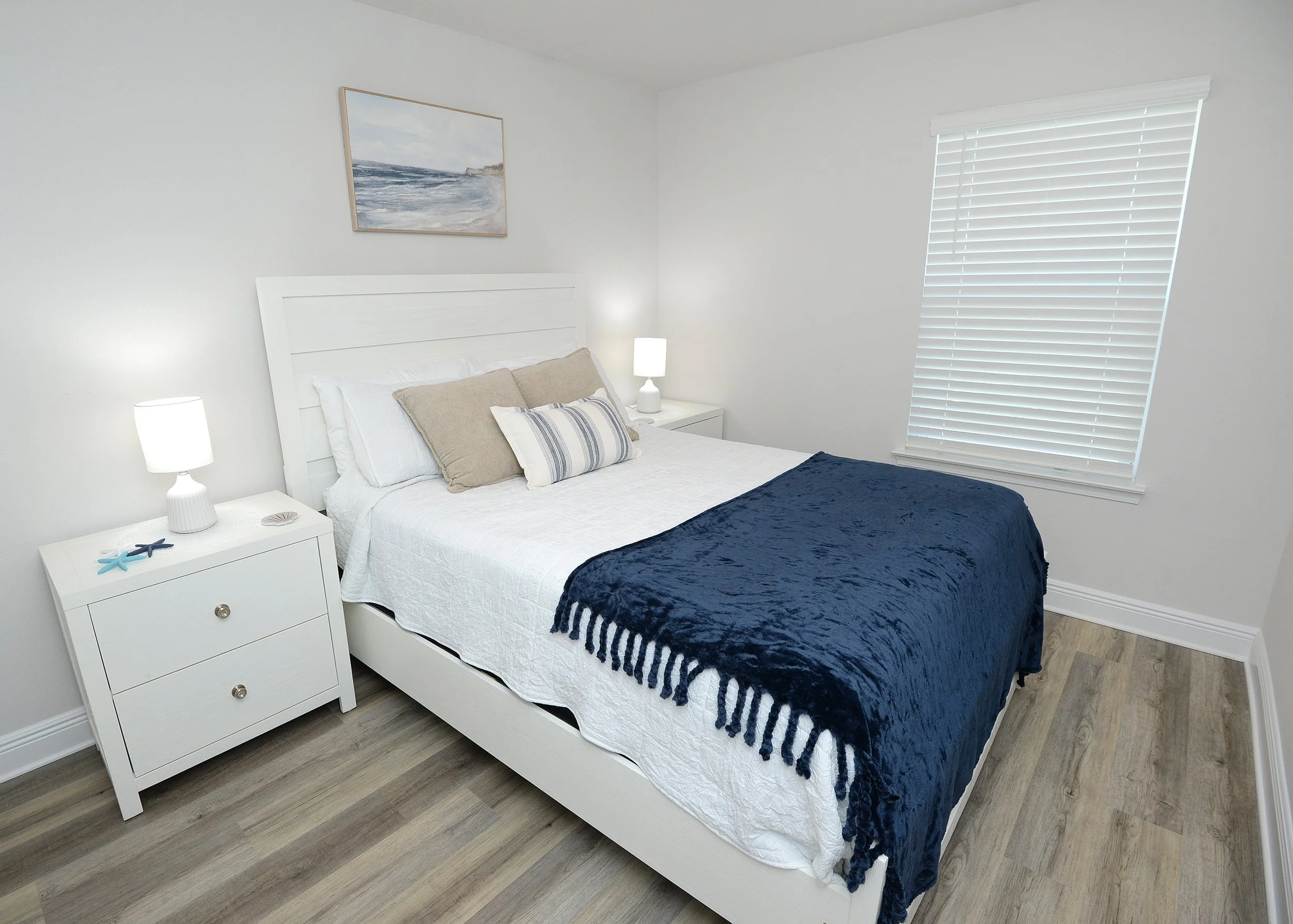 A bedroom with white walls and a white bed frame. The bed is made with white bedding, beige and striped pillows, and a dark blue quilt. There are two white nightstands with lamps on each side of the bed. A painting of an ocean scene hangs above the b