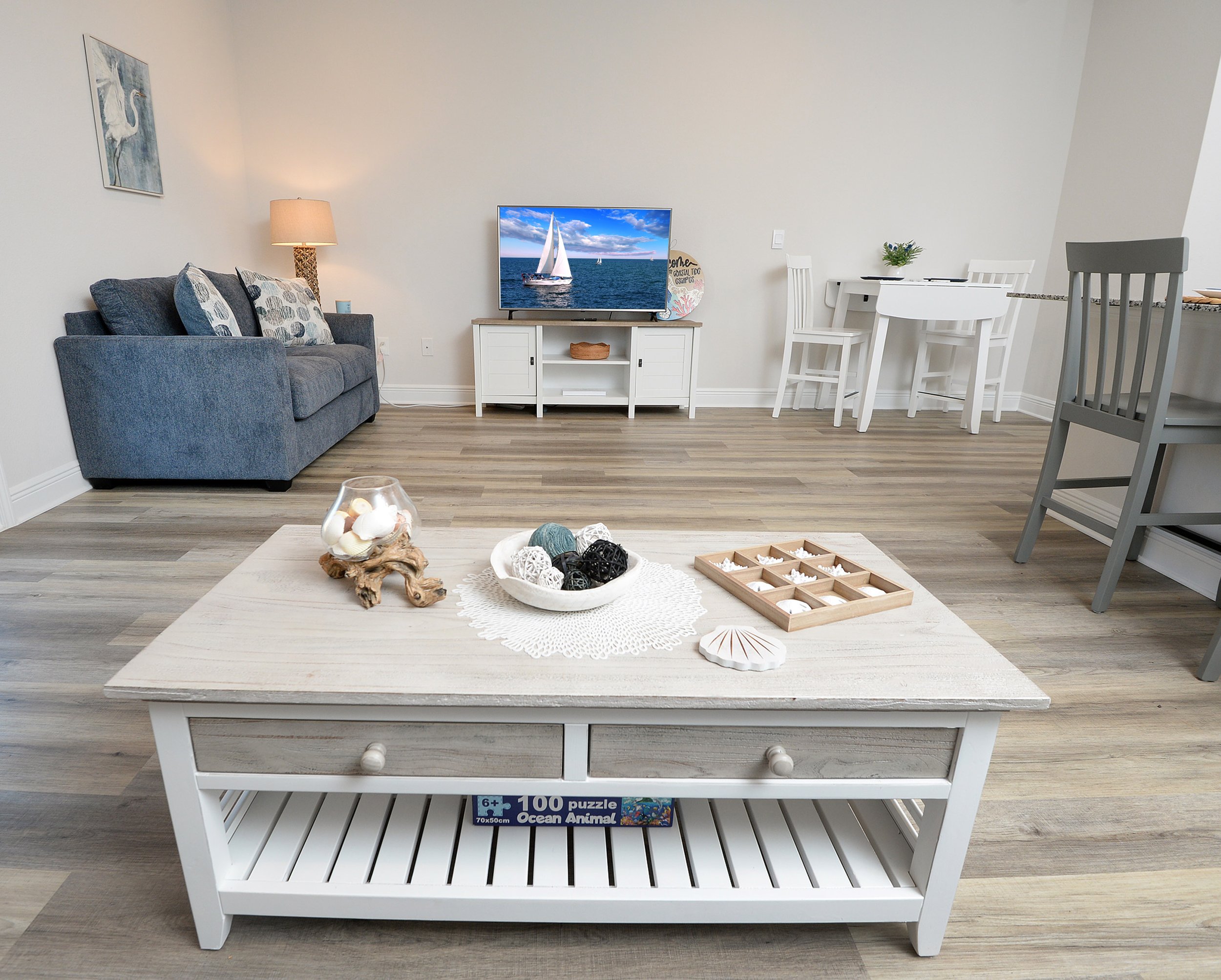 A living room with a gray sofa, a white coffee table, a TV on a white stand, and a dining table with chairs. There is wall decor, a lamp, and a coffee table with decorative items.