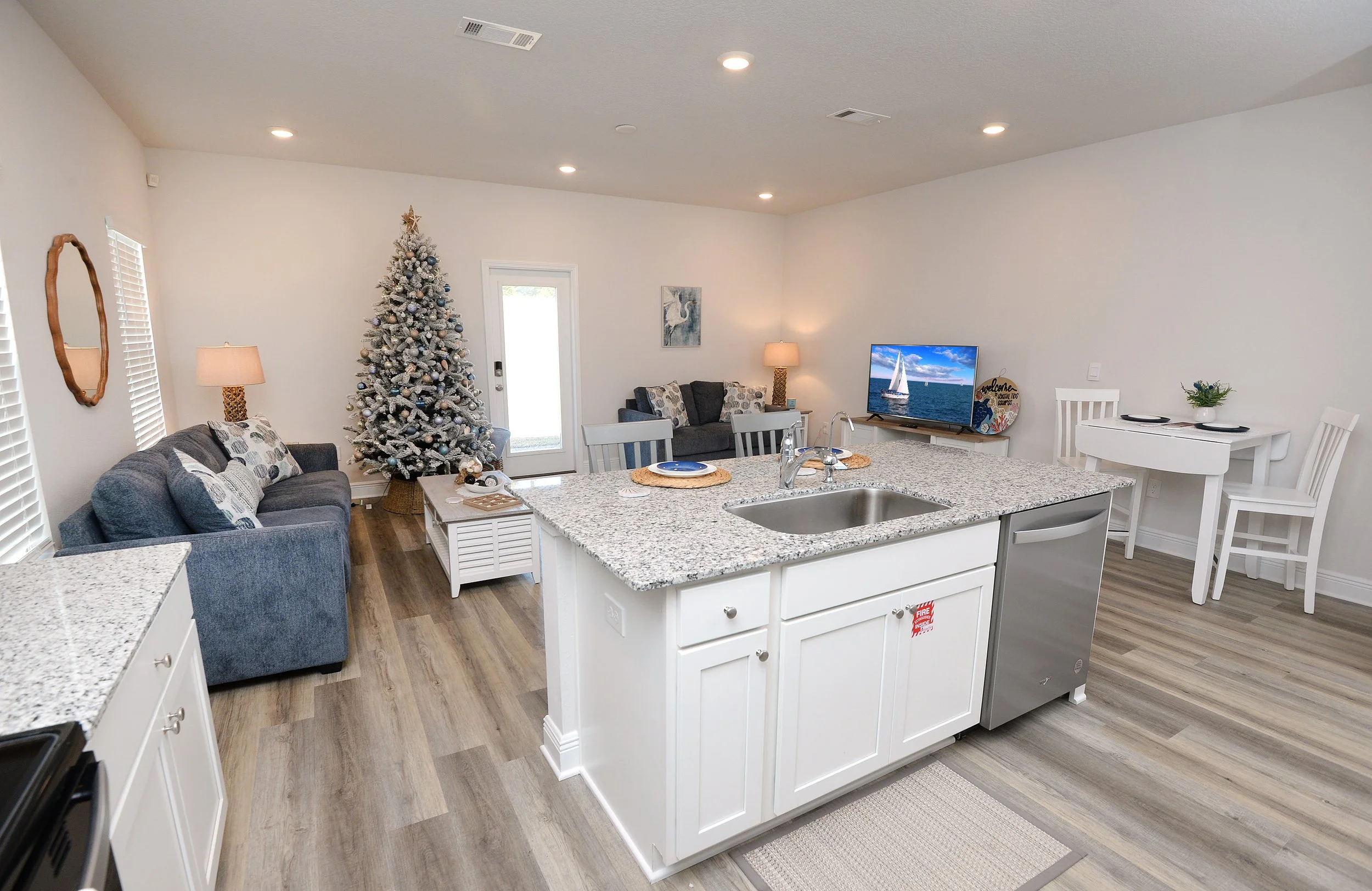 A living room with a Christmas tree decorated with blue and silver ornaments, a gray sofa, a coffee table, two lamps, a wall art of swans, a TV showing a sailboat, and a small white dining table with chairs.