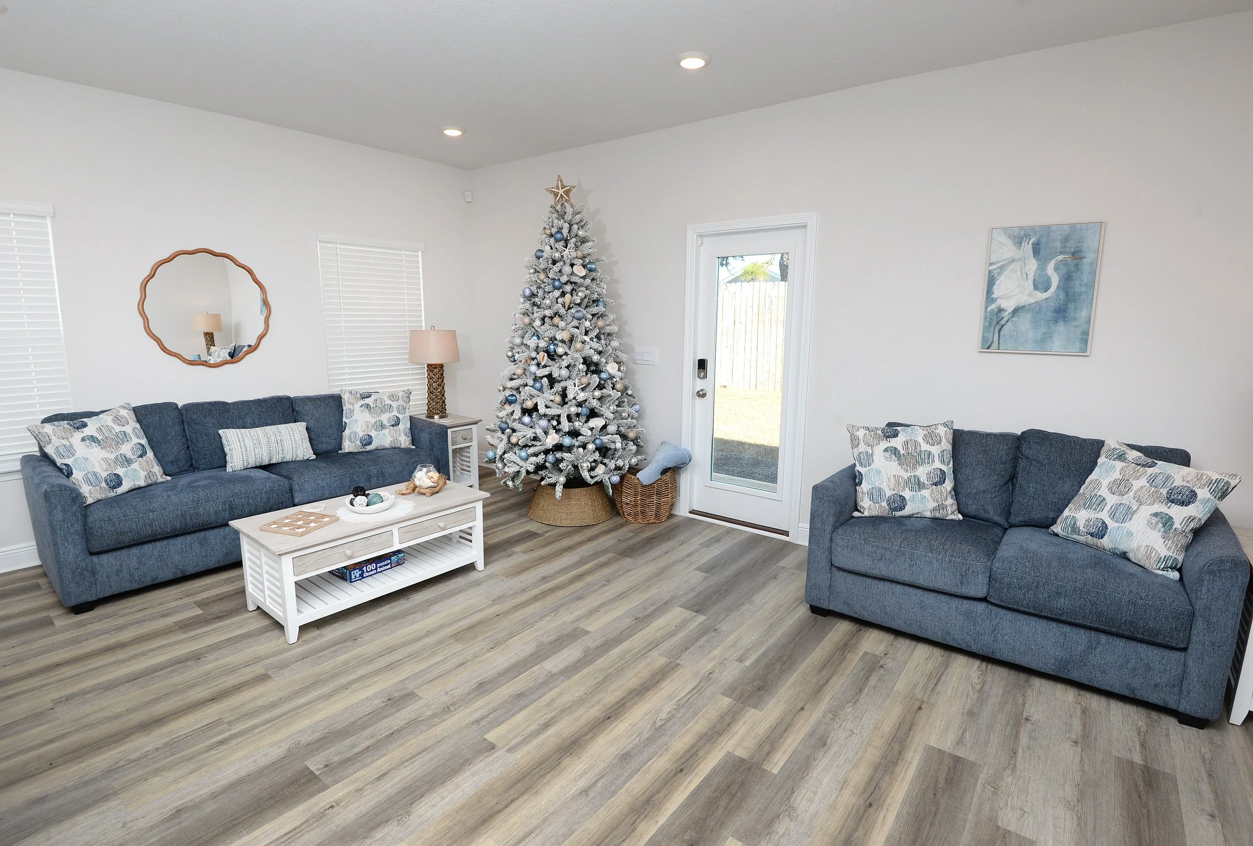 Living room with two gray sofas and throw pillows, a white coffee table with a puzzle box, a decorated Christmas tree, a mirror, a floor lamp, and a wall art of a heron, with a door leading outside.