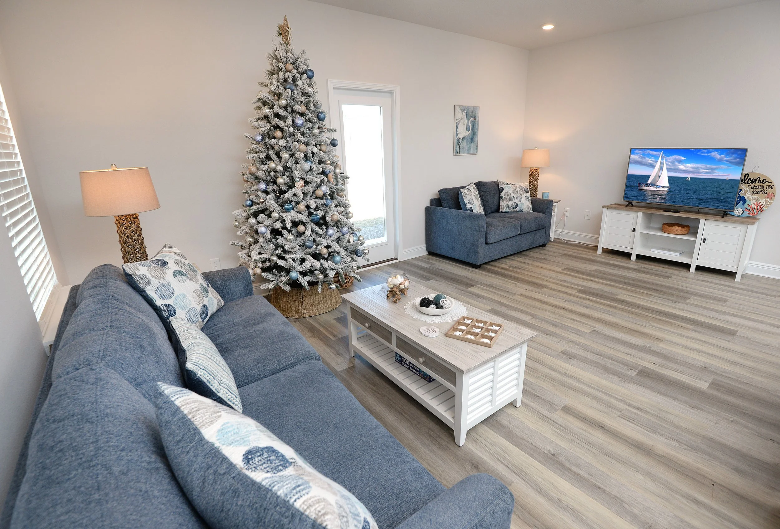 A living room decorated for Christmas with a white flocked Christmas tree, a blue couch with patterned pillows, a coffee table, and a TV on a white stand displaying a sailboat on the water.