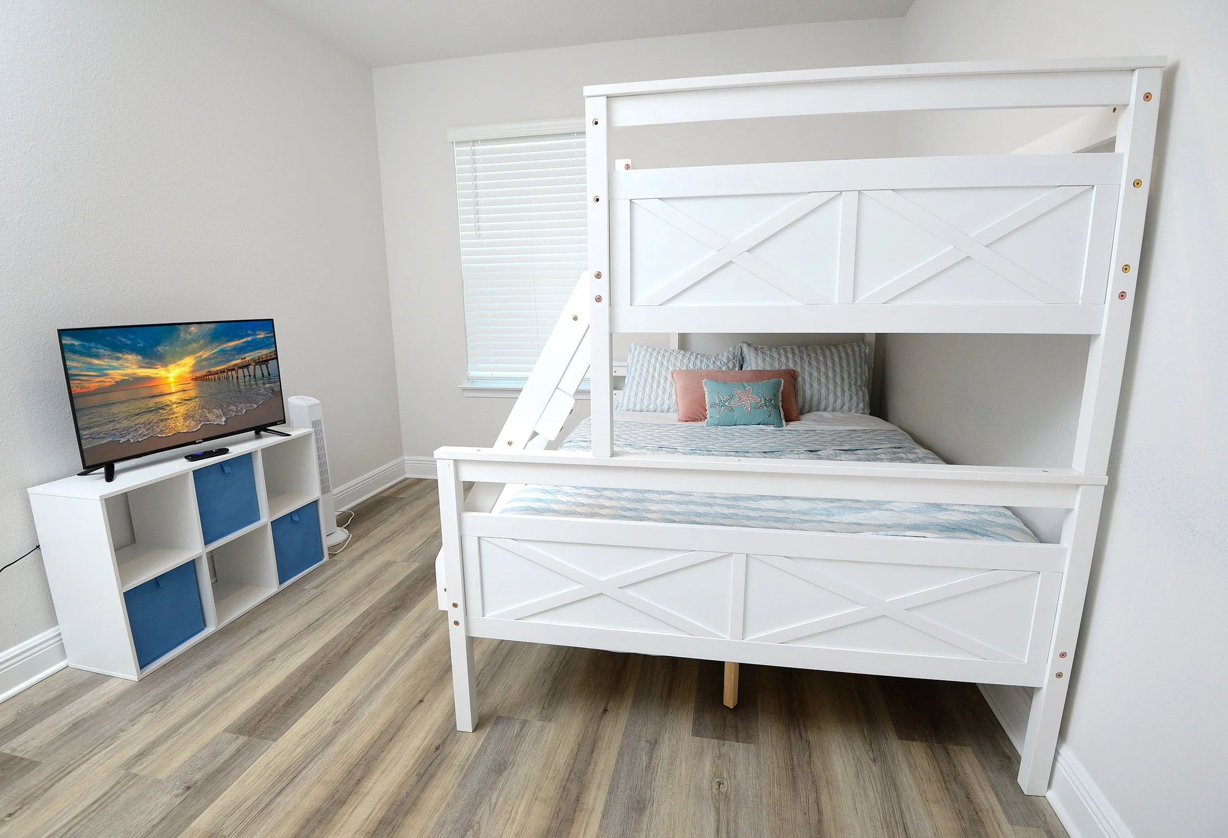 A bedroom with a white bunk bed, a small TV on a white and blue cubical storage unit, and a window with horizontal blinds. The room has light-colored wood flooring and white walls.