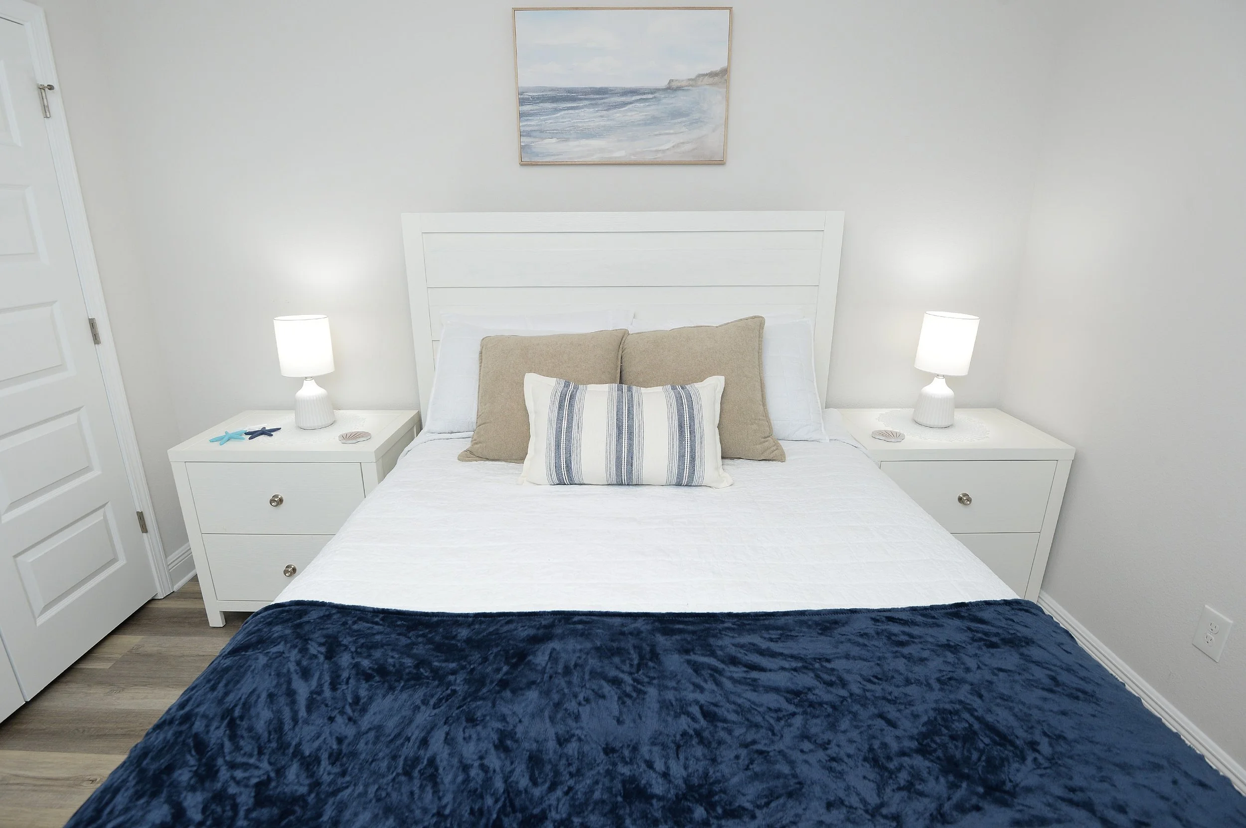 A bedroom with a white bed, beige pillows, and a navy blue blanket at the foot of the bed. Two white nightstands with lamps, starfish, and seashell decorations. A landscape painting of a shoreline with cliffs hangs above the headboard. White walls an