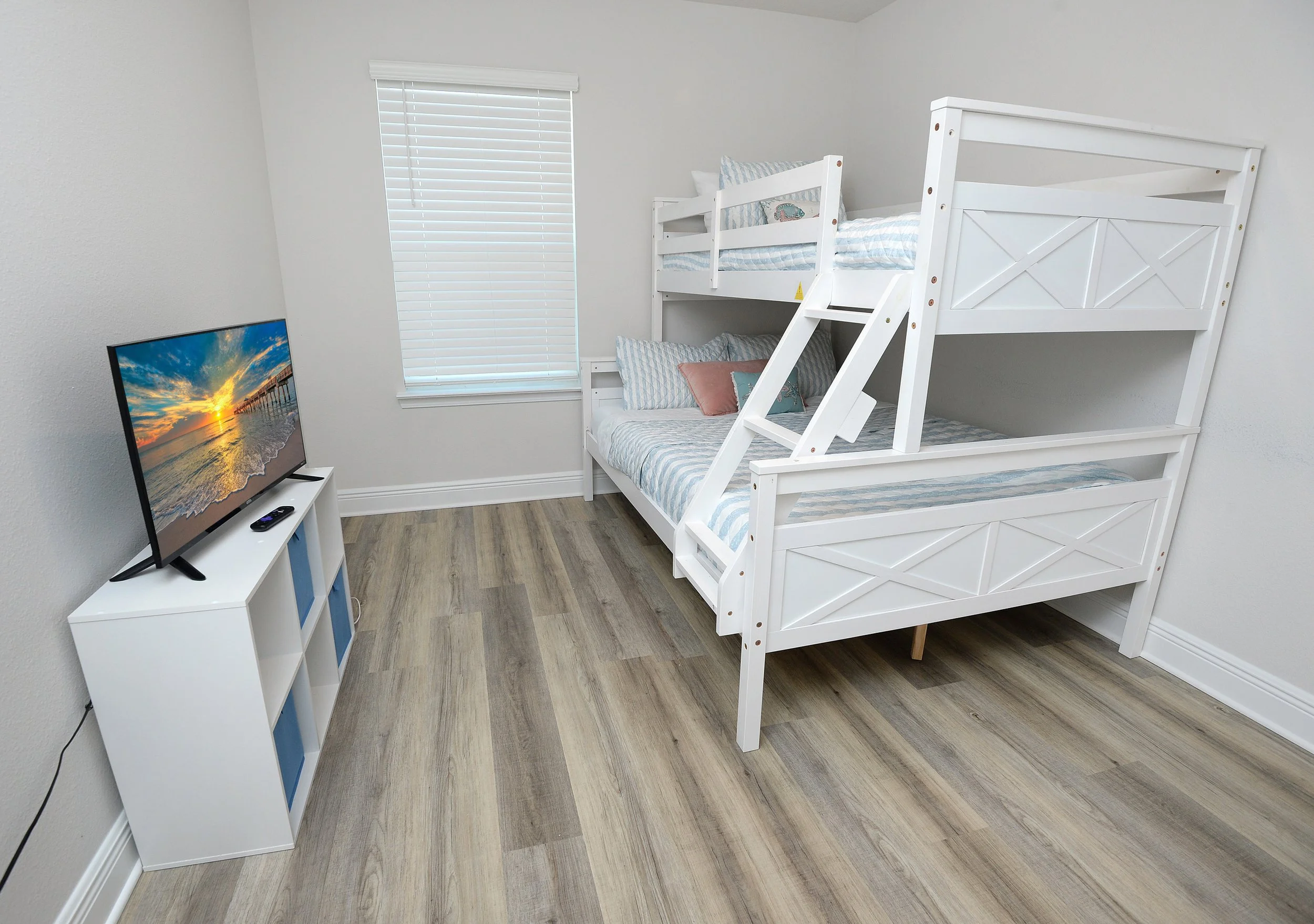 A bedroom with white walls and a wood floor. There is a bunk bed with white wood and blue and white bedding. A small white storage unit with blue baskets holds a flat-screen TV displaying a sunset scene. There is a window with closed white blinds.