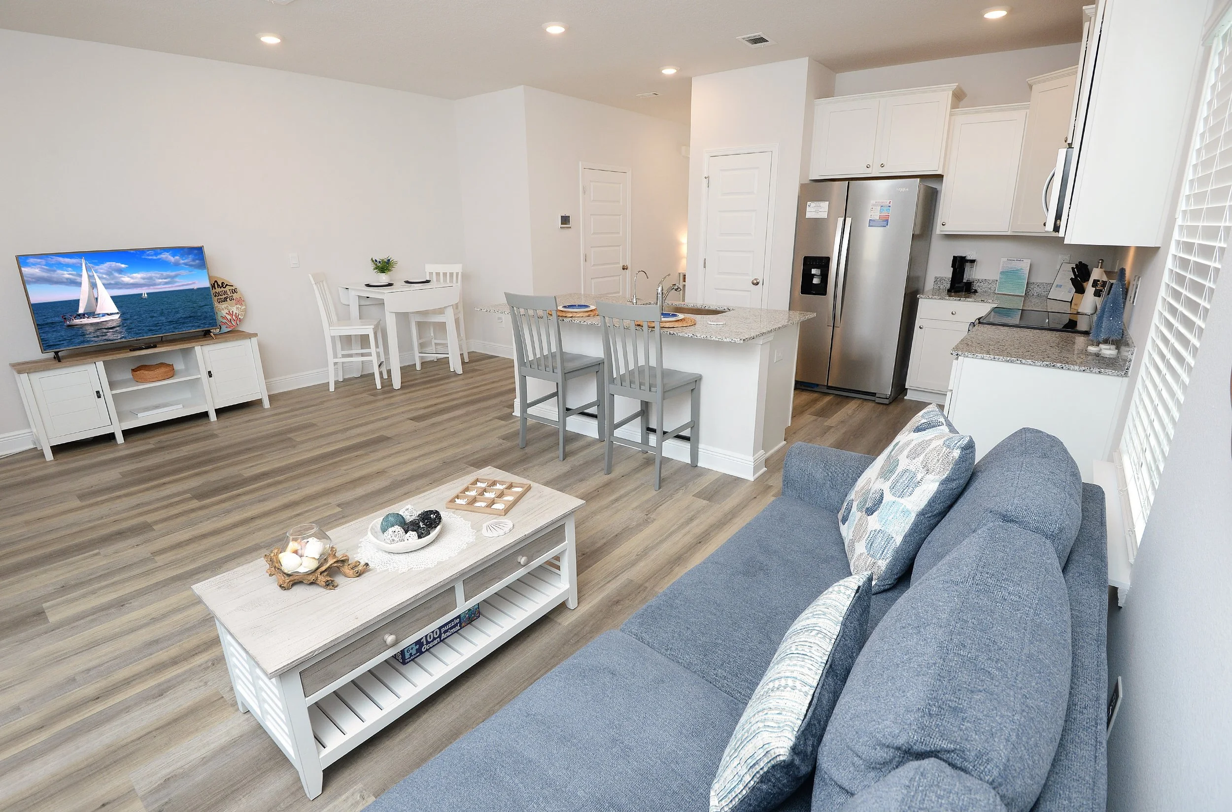 Living room with a blue couch, a white coffee table with decorative objects, a TV on a stand, a small dining table with two chairs, a kitchen with white cabinets, stainless steel refrigerator, and granite countertops. There is a window with blinds an