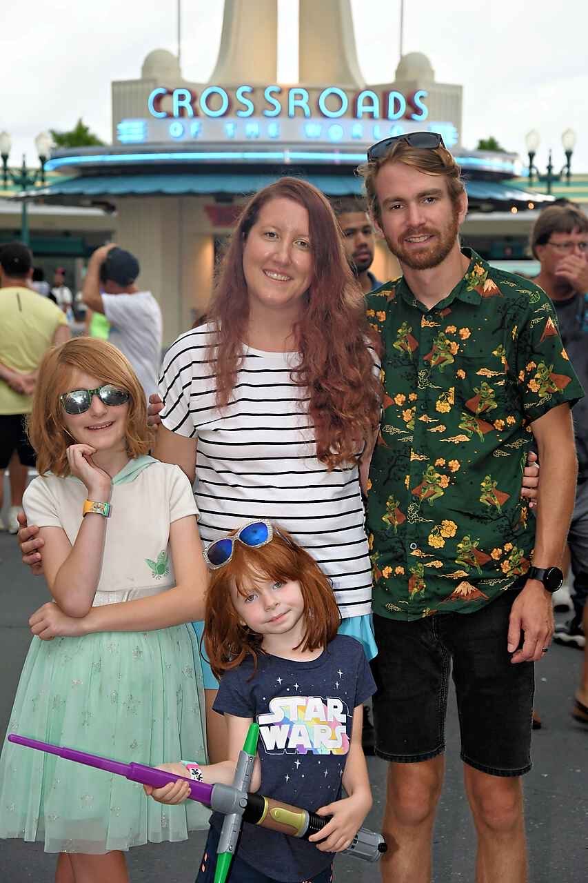 A family of four posing in front of the Crossroads of the World sign at a theme park. The mother has long red hair and is wearing a striped shirt. The father has short light brown hair, a beard, and is wearing a tropical shirt. The daughter has shoulder-length red hair, sunglasses, and a butterfly shirt. The younger girl has red hair, a Star Wars shirt, and is holding a toy lightsaber.