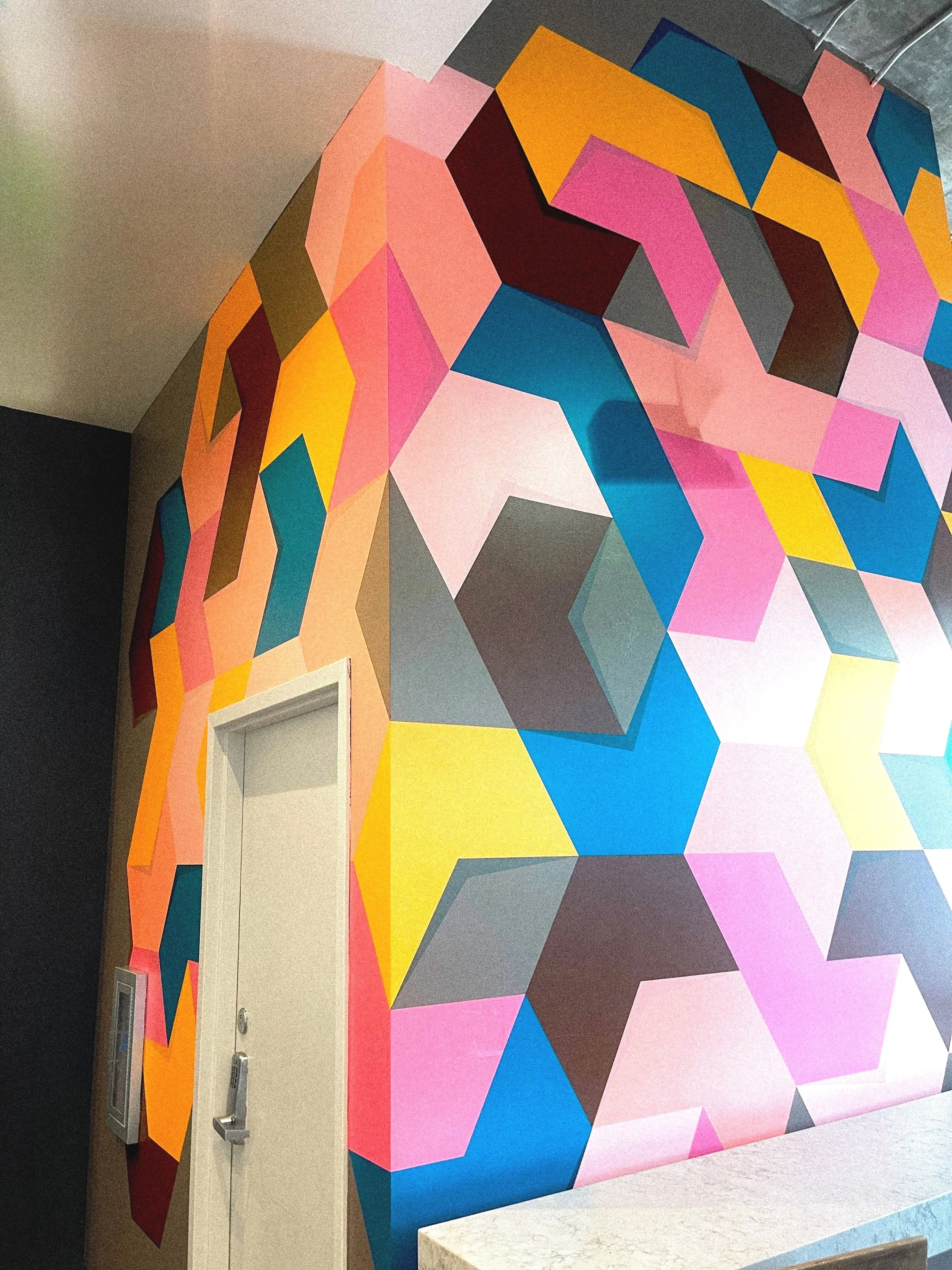 Colorful geometric mural painting on a wall with shades of pink, purple, blue, yellow, orange, and black.