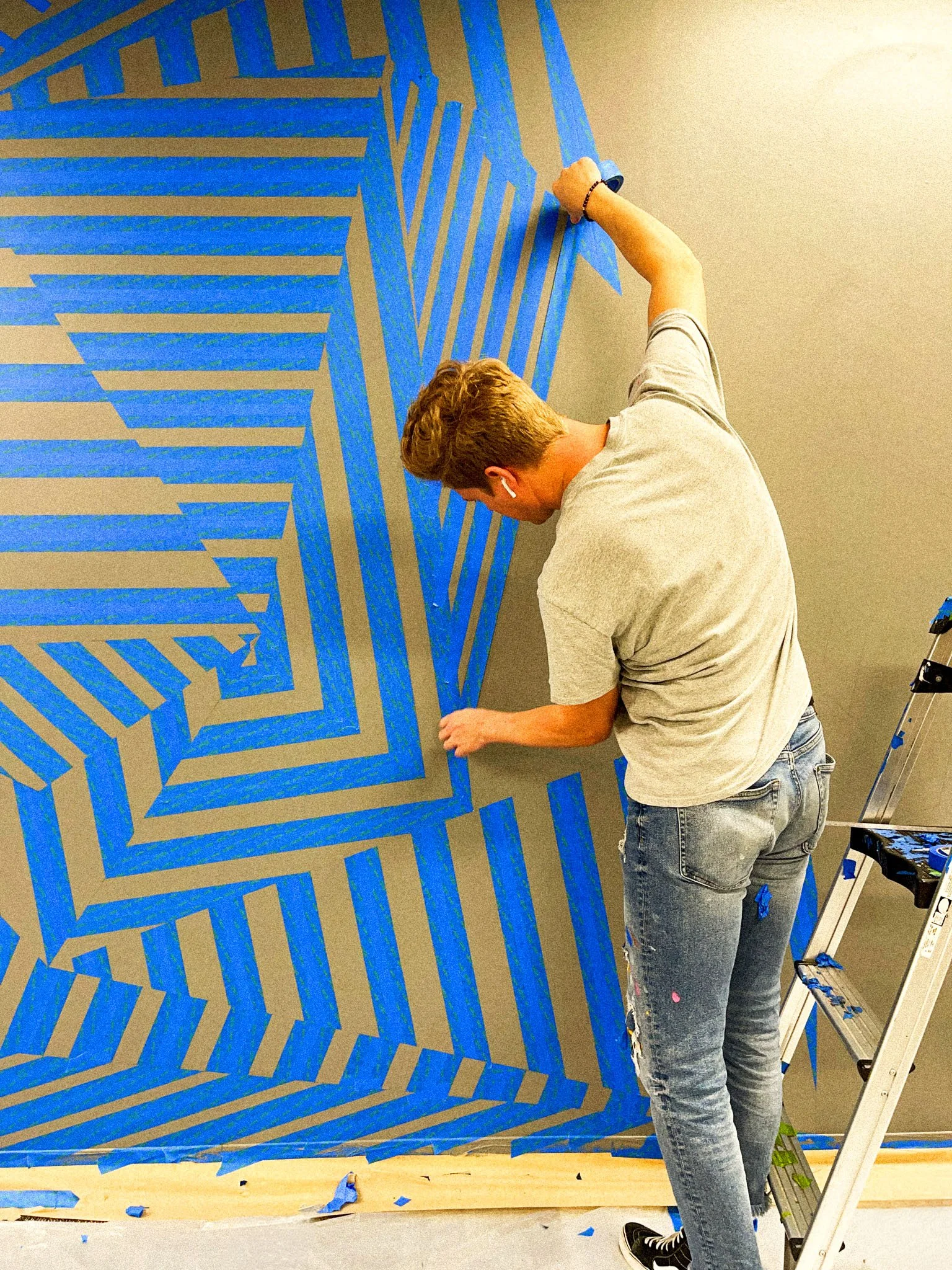 Andrew Haan installing a geometric office mural in Laguna, California, using blue painter’s tape to map the angular striped design.