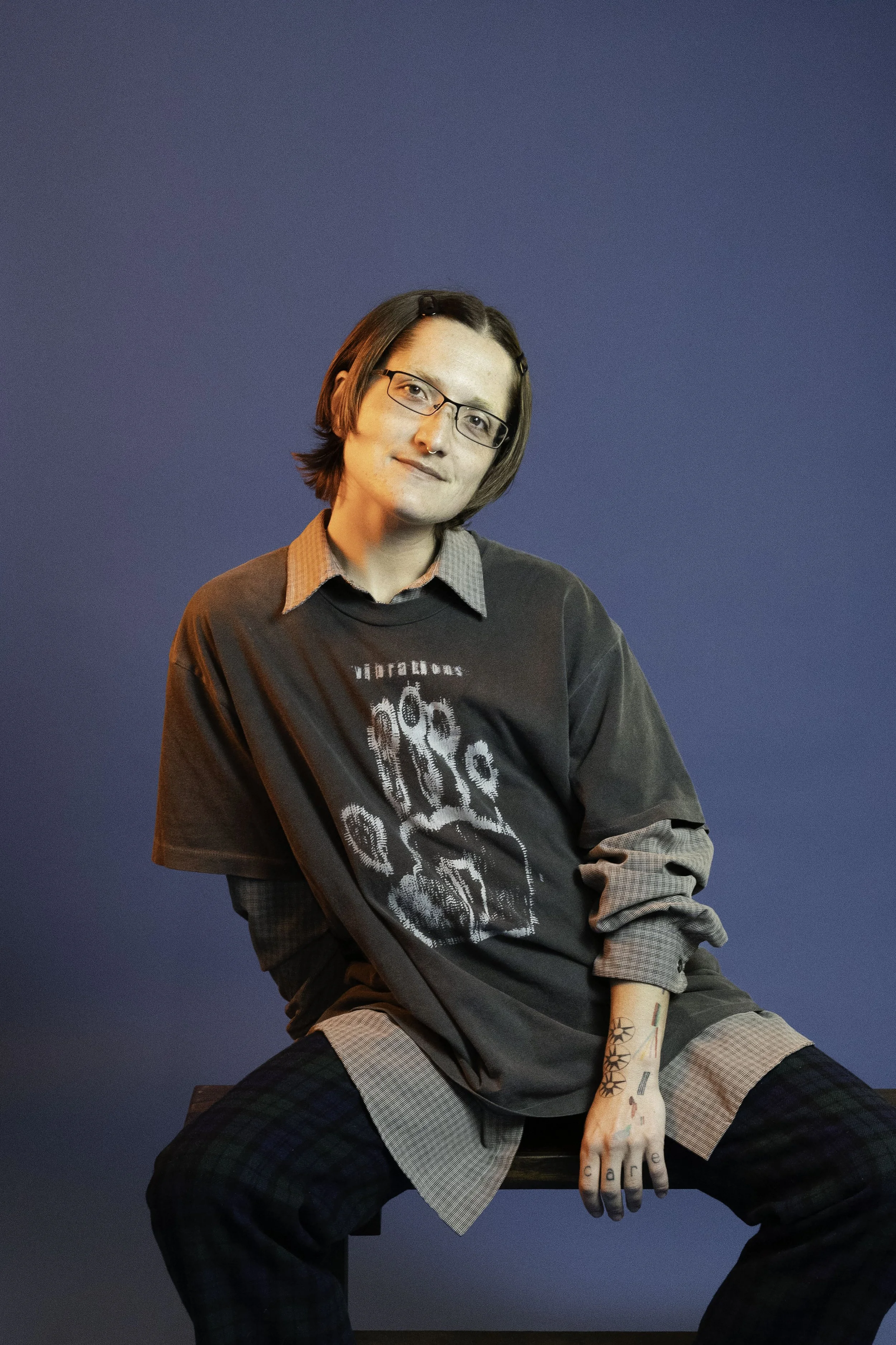 A young person with shoulder-length brown hair, glasses, and tattoos on their right arm sitting on a wooden chair against a blue background. They wear a black graphic t-shirt, a plaid skirt, and a checkered shirt layered underneath, with a slight smile and relaxed pose.