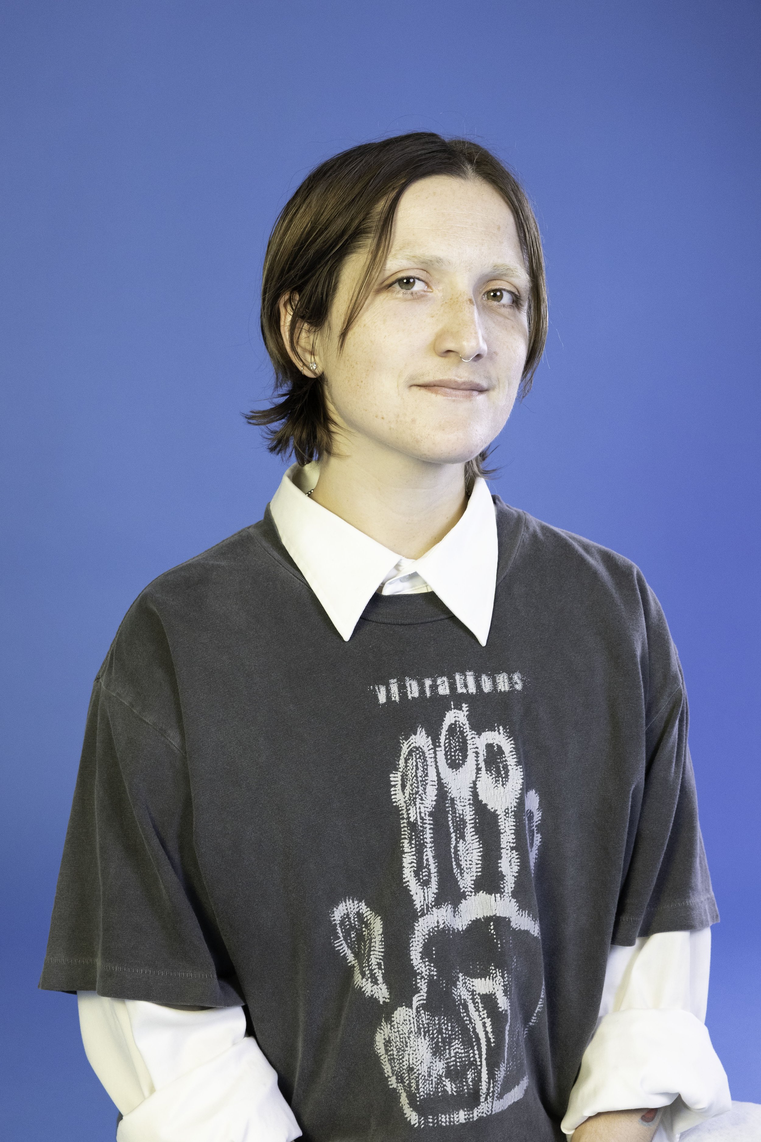 A person with short brown hair, wearing a white shirt layered with a dark gray graphic t-shirt, and a septum piercing, standing against a solid blue background.