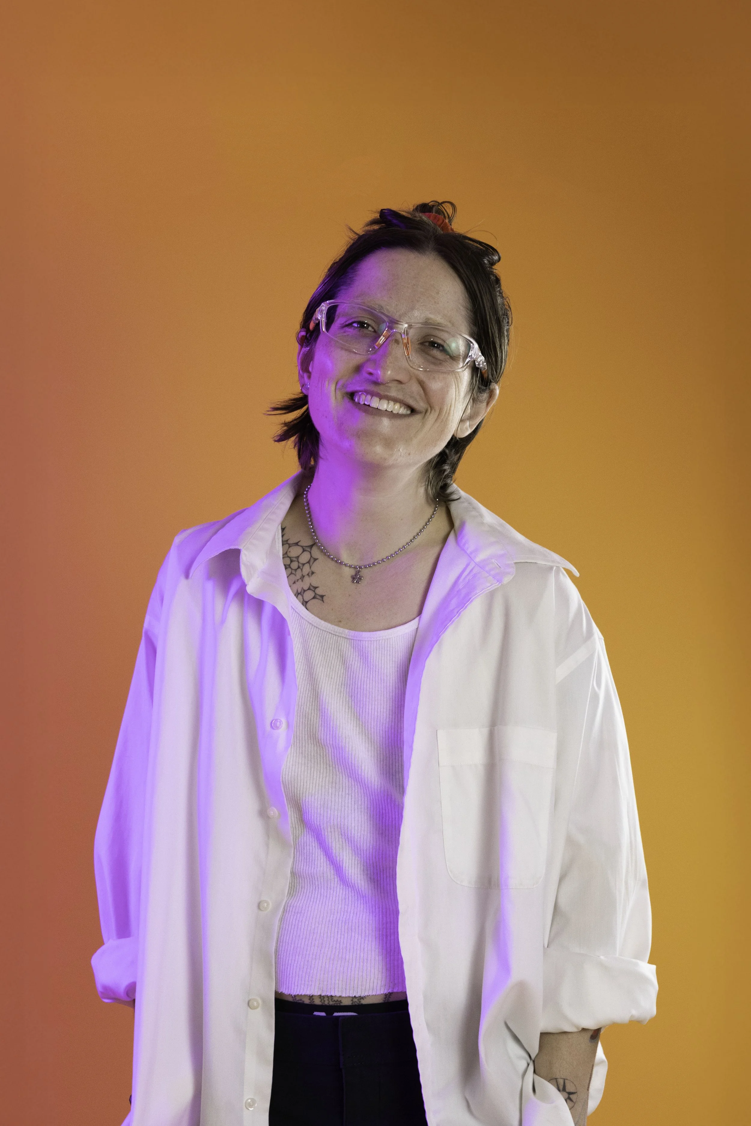 Person with short dark hair, glasses, and tattoos, smiling, wearing a white shirt and standing against a bright orange background with purple lighting.