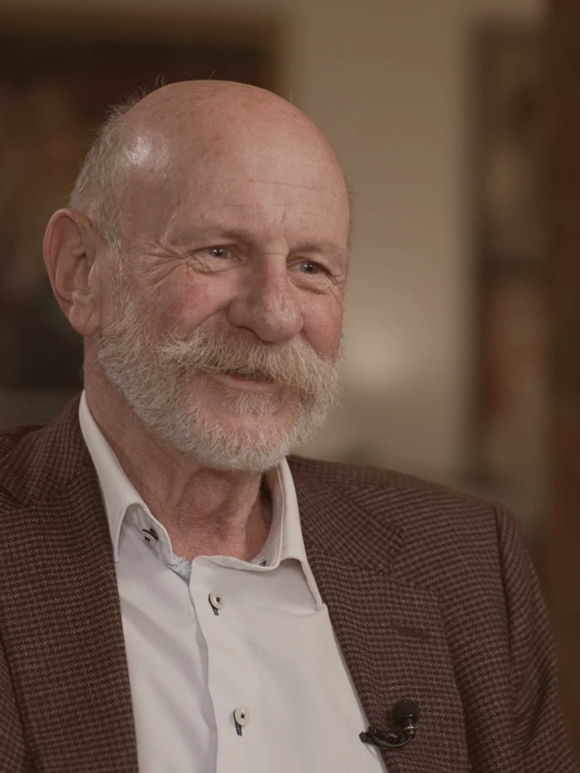 Mark Sainsbury, renowned and beloved NZ Kiwi interviewer and storyteller smiles at his interviewee positioned off camera. Mark wears a patterned brown suit jacket with a cream shirt.