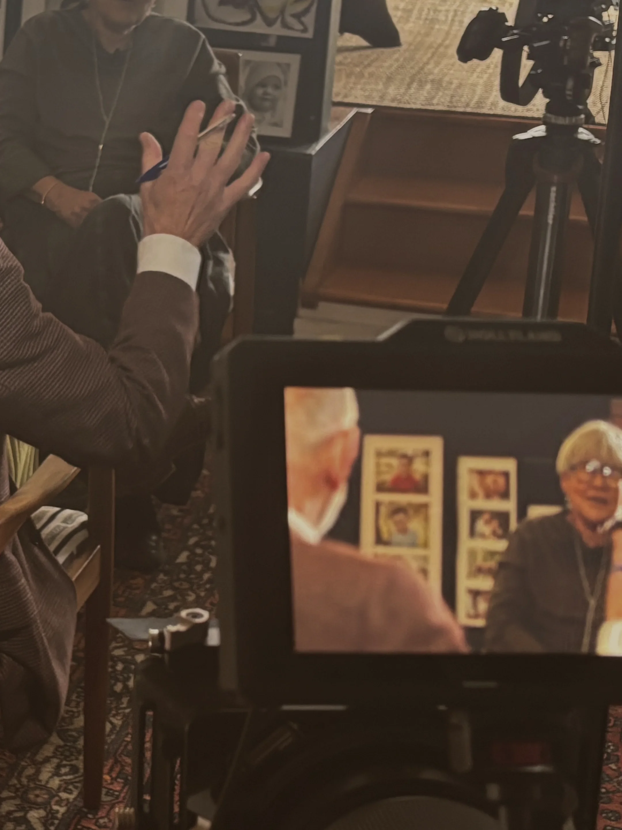 A close-up of a professional video camera, focussed on an elderly woman being interviewed by elderly man in a comfortable grandmother's living room.
