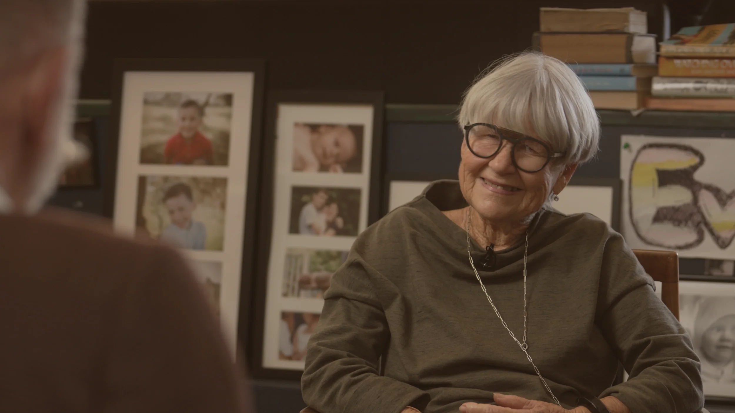 Elderly woman with short bob hairstyle and big glasses is interview by Mark Sainsbury in her comfortable living room.