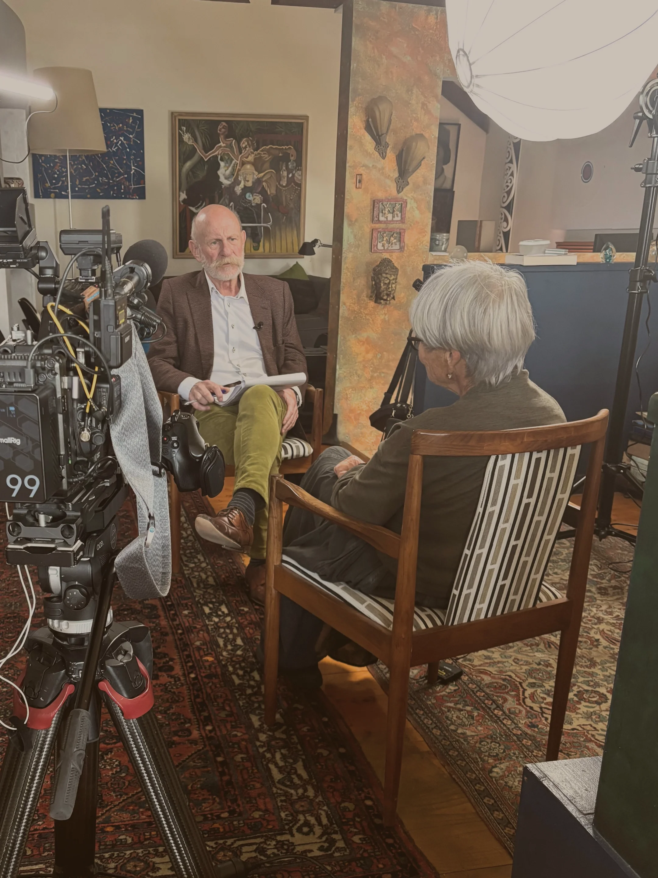 Elderly man and renowned NZ storyteller, Mark Sainsbury, interviews an elderly woman about her life, on camera with professional cinematography equipment and crew, set up in a grandmother's comfortable living room.