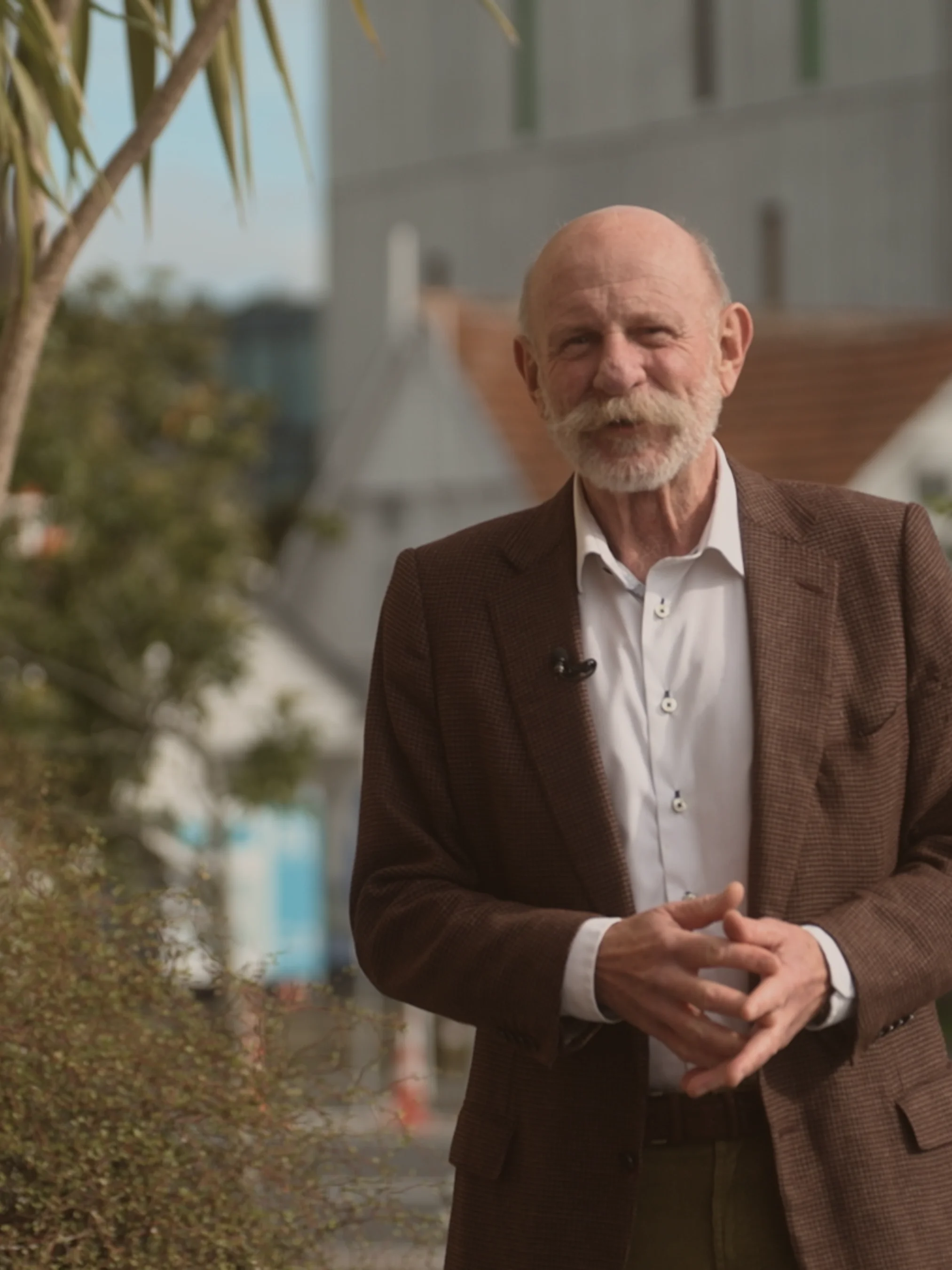 An elderly man wearing a brown corduroy jacket, the renowned Kiwi television personality, Mark Sainsbury, speaks in front native plants on a suburban street.