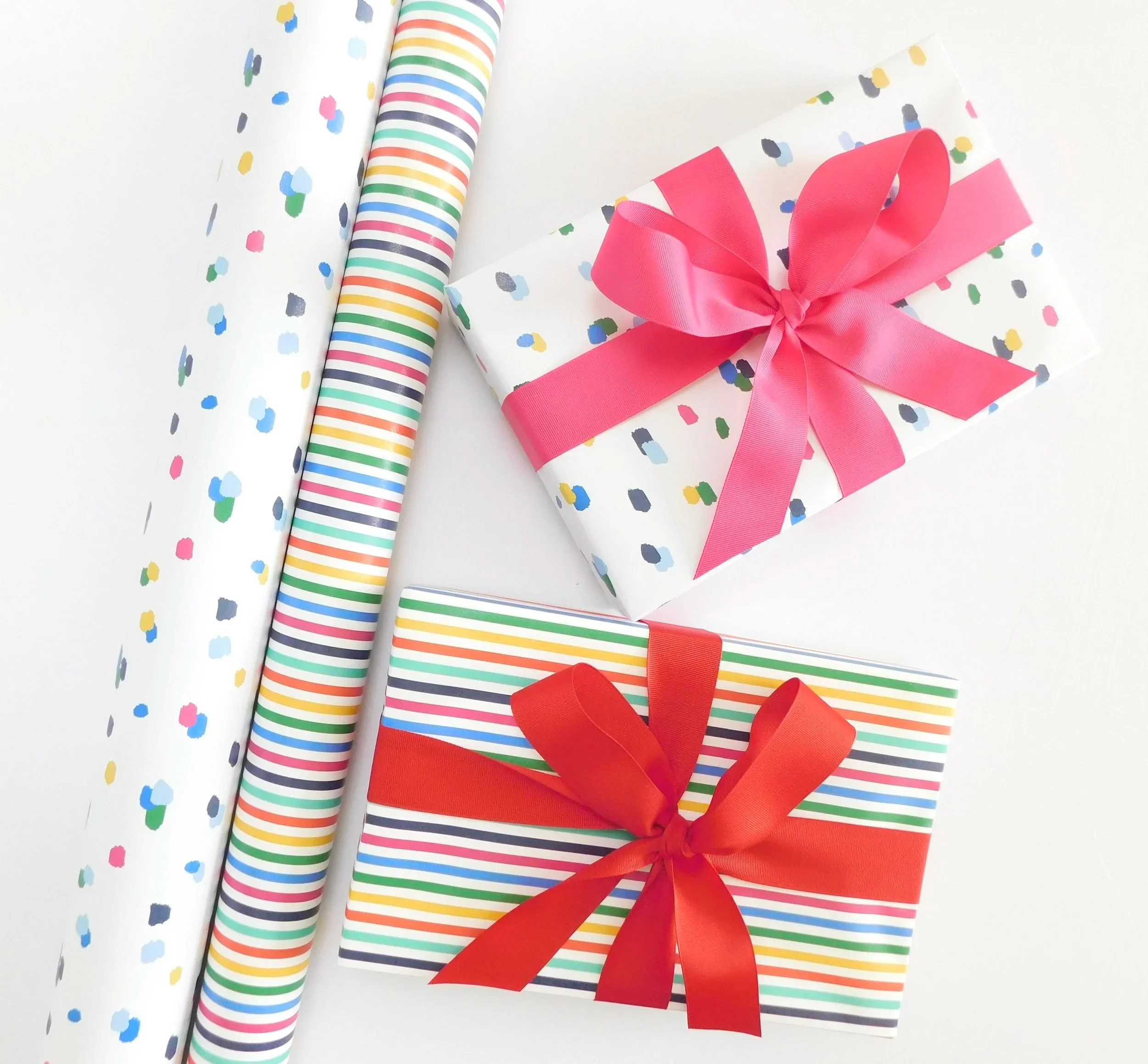 A gift wrapped in white paper with multicolored confetti-like dots, decorated with a large pink ribbon bow, along with a roll of confetti-themed wrapping paper with multicolored stripes and confetti on a white surface.