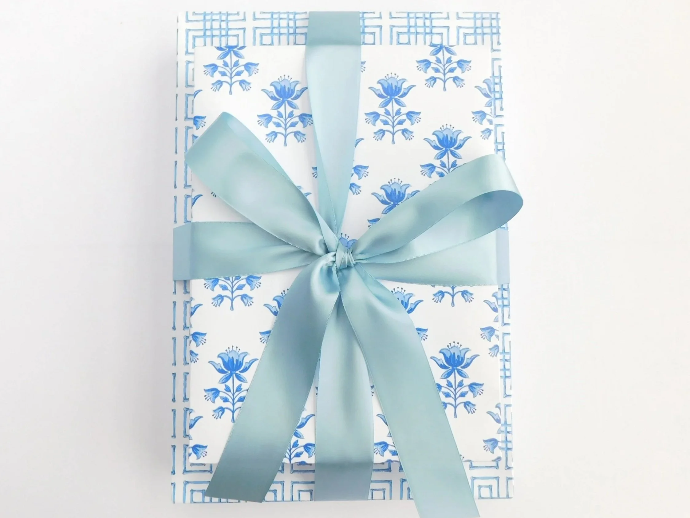 A gift box wrapped in white paper with blue floral patterns, tied with a wide light blue ribbon tied into a bow.