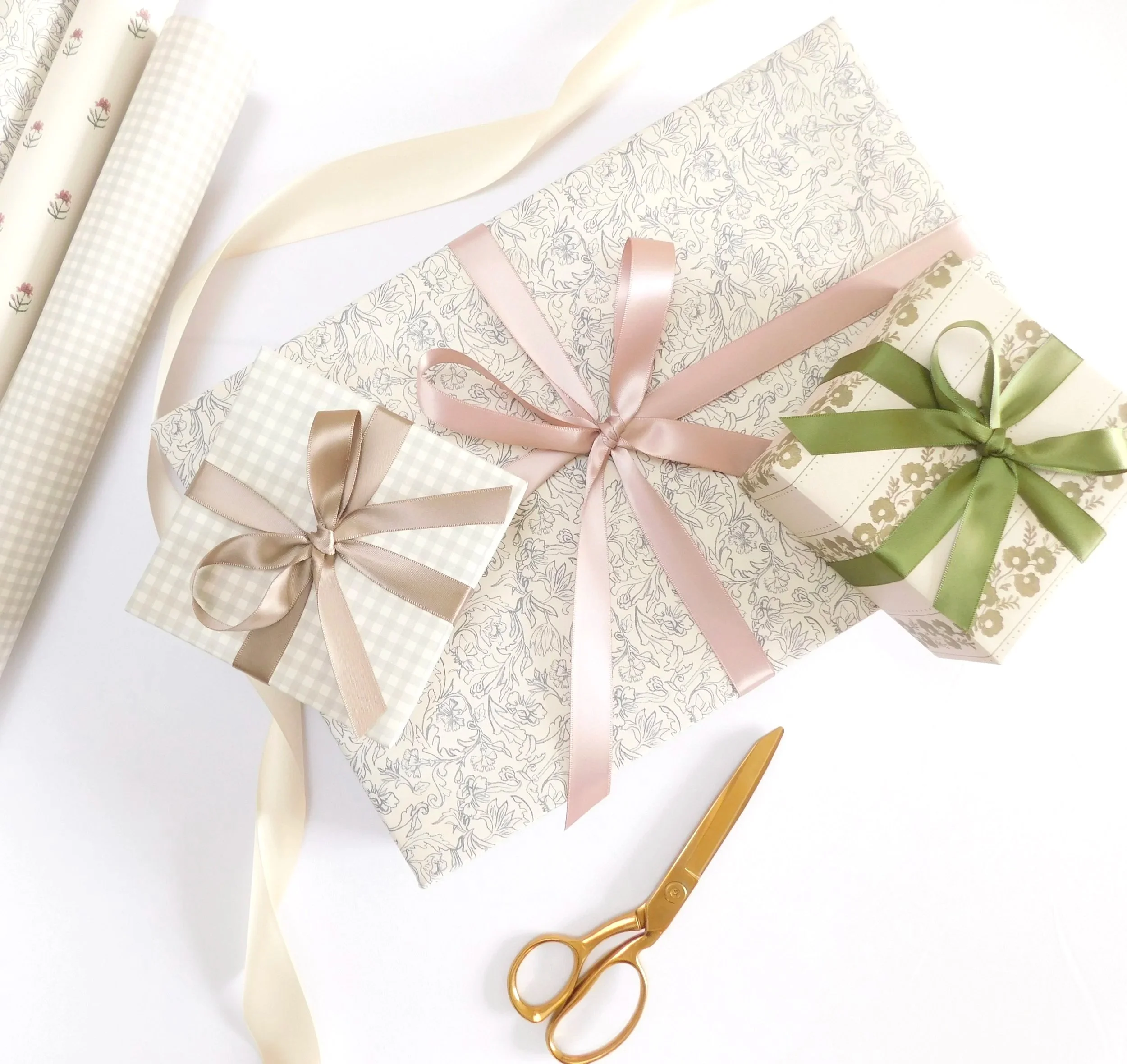 Various rolls of patterned and plain wrapping paper, some with floral and checkered designs, arranged on a white surface. Ribbons and a pair of gold scissors are also present.