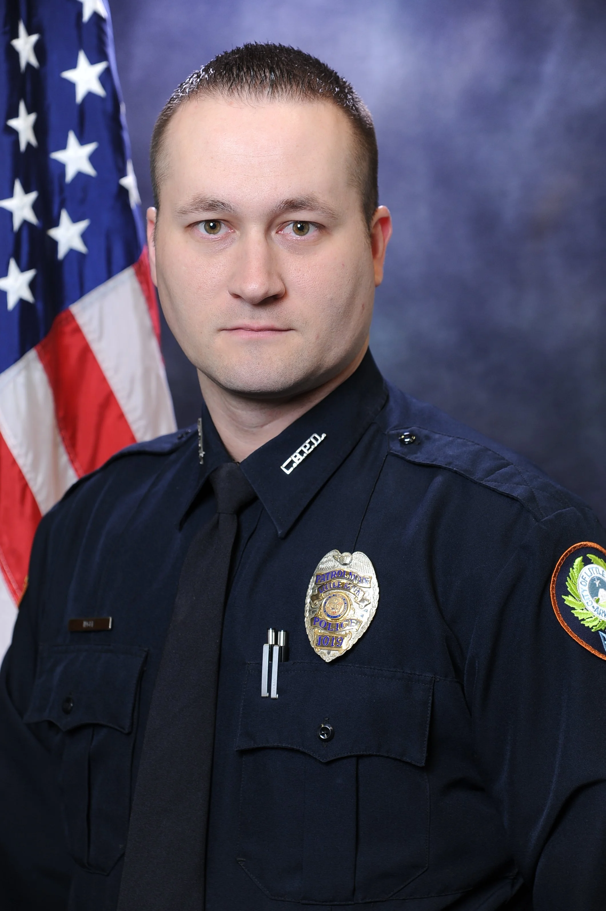 A police officer in uniform standing in front of an American flag and a blue background.