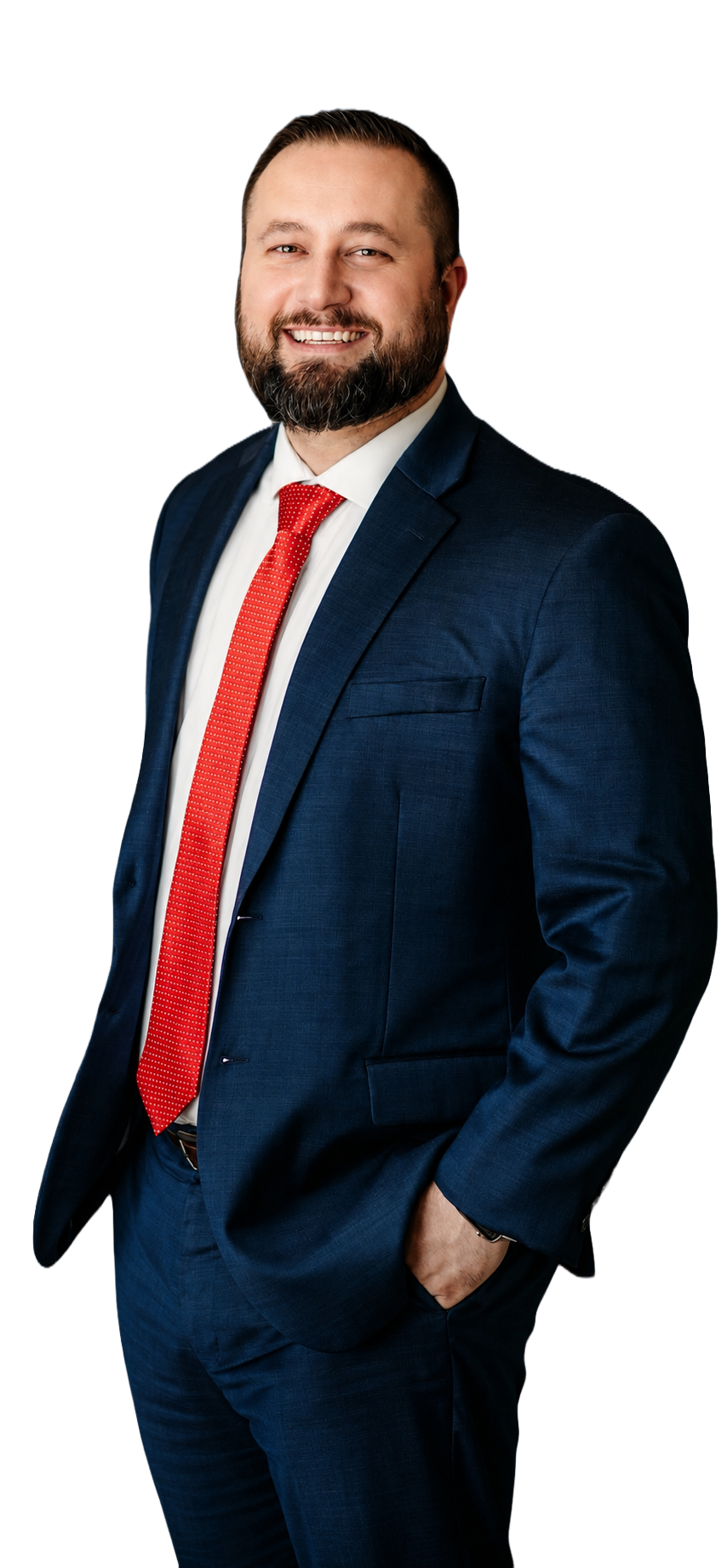 A smiling man in a navy blue suit, white shirt, and red tie, standing with his right hand in his pocket against a black background.