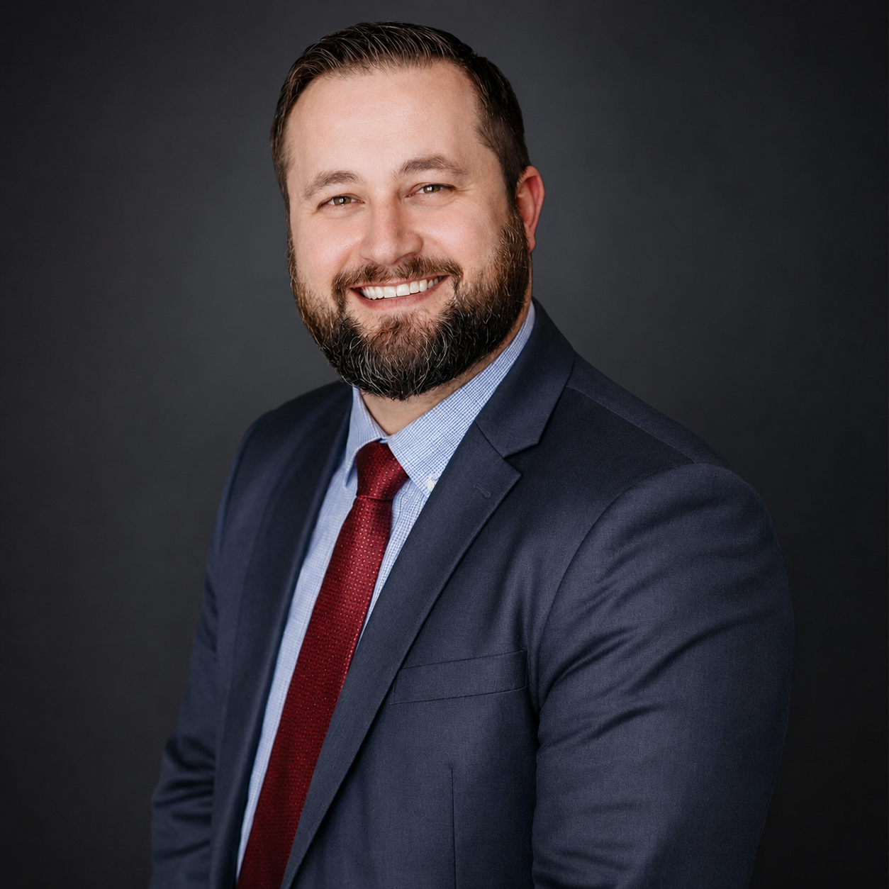 Professional headshot of a man with a beard in a dark suit, light blue shirt, and red tie, smiling against a dark background.