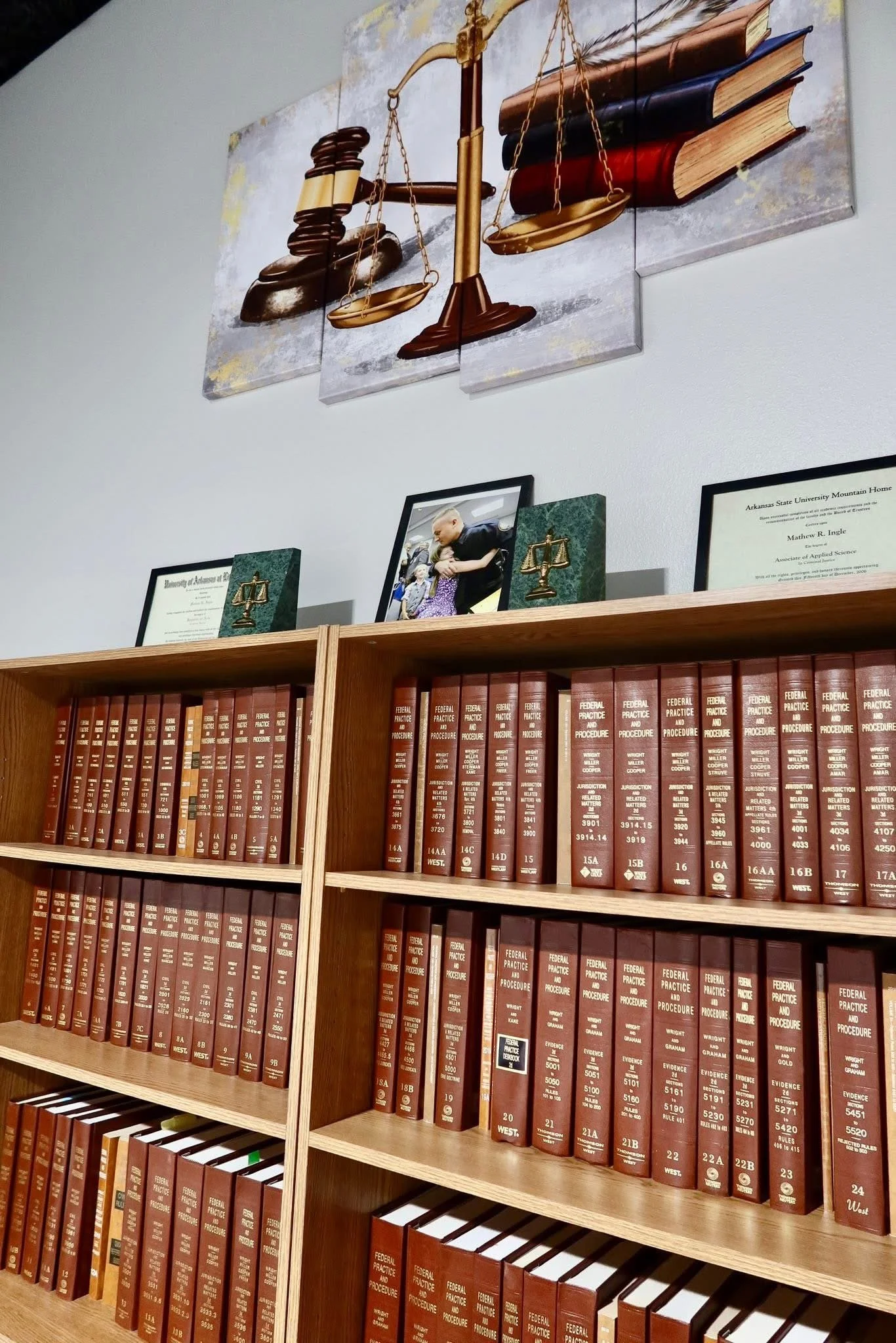 Wooden bookshelf filled with law books and framed documents, with a wall art featuring a gavel, scales, and books above the shelf.