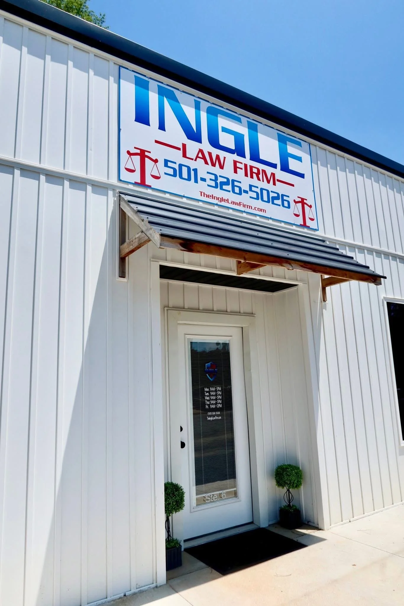 Exterior of a white law firm office with a sign displaying the firm's name, phone number, and website. The entrance has a glass door with small potted plants on either side and a black doormat.