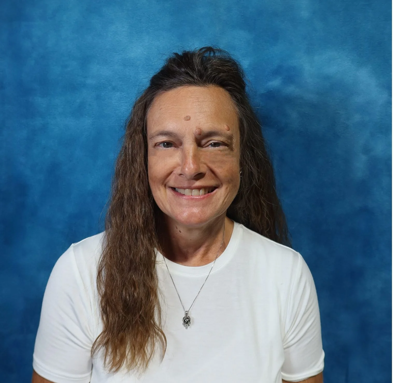 A woman with long, wavy brown hair, wearing a white shirt and silver necklace, smiling against a blue textured background.