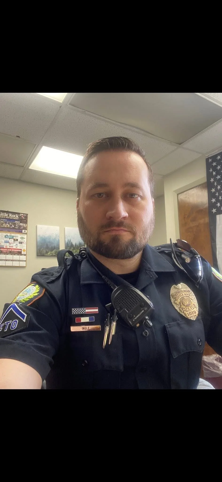 A police officer taking a selfie in uniform inside a room, with an American flag and a sheriff badge visible.