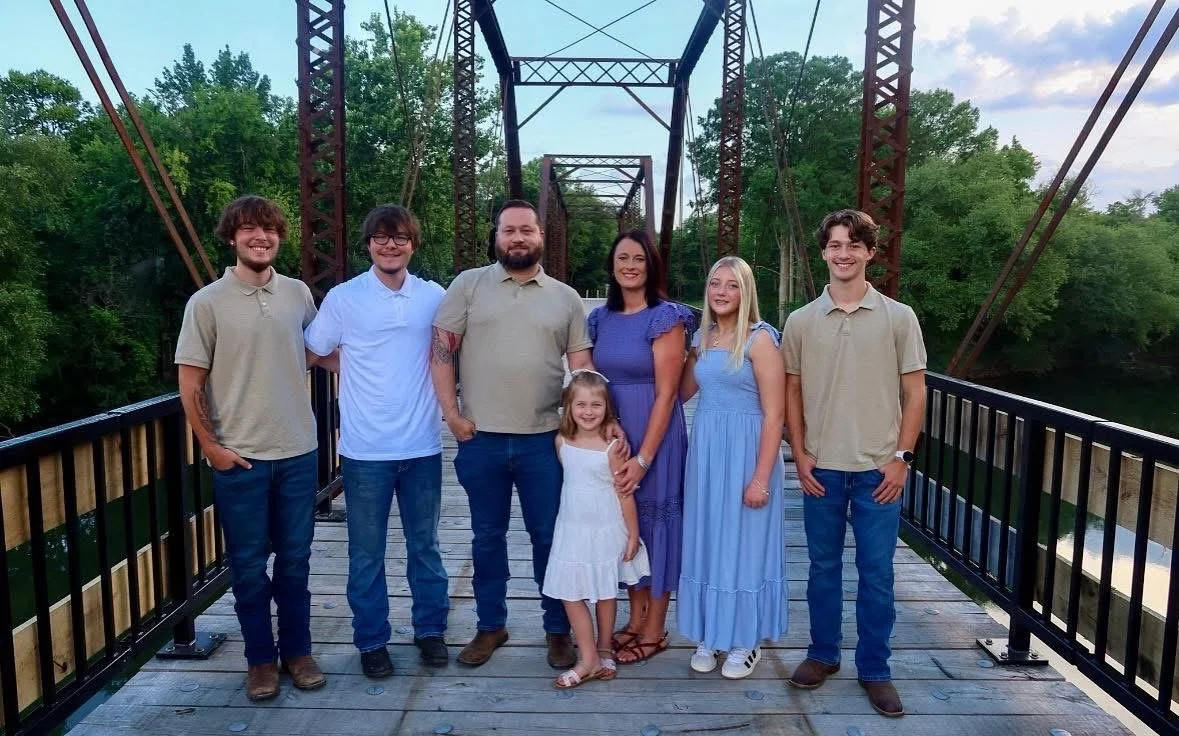 Family photo of seven people standing on a wooden bridge surrounded by trees
