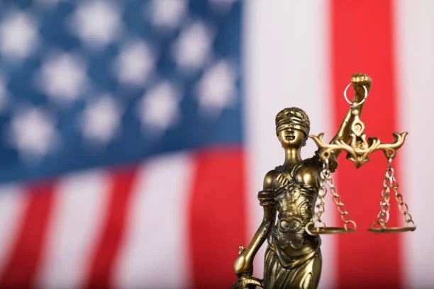 Close-up of a golden statue of Lady Justice holding scales, with an American flag in the background.