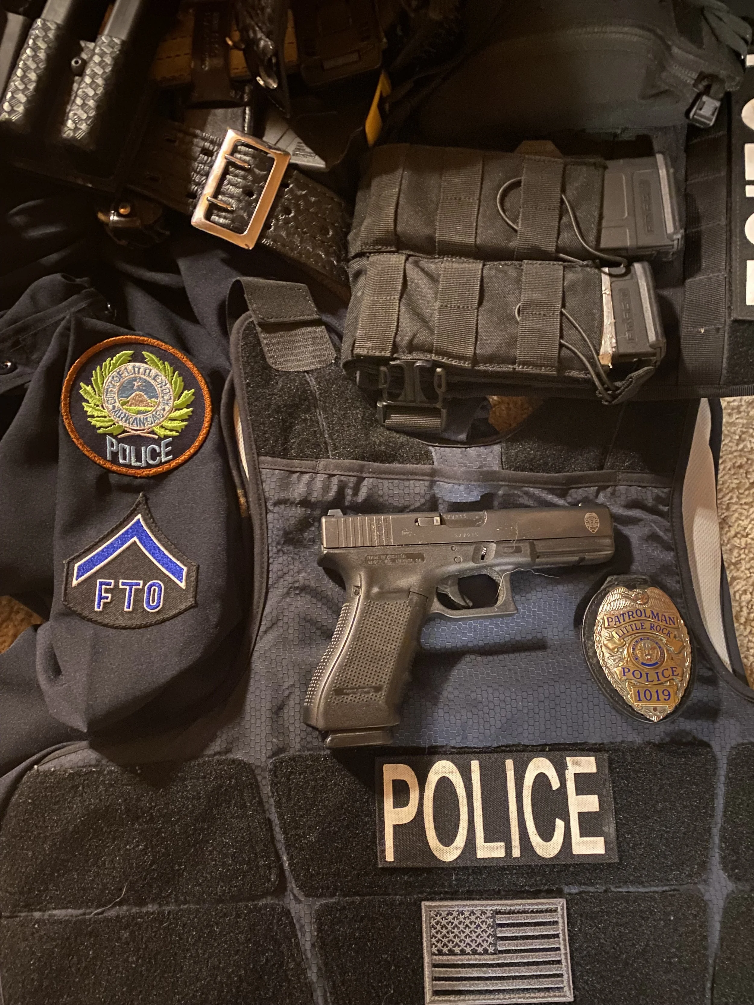 Police badge, firearm, tactical gear, and police patches laid out on a black surface.