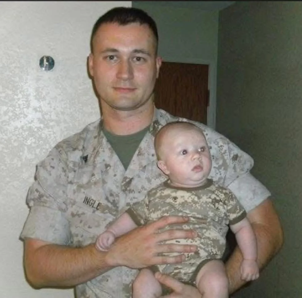 A man in military uniform holding a baby also dressed in camouflage, standing indoors against a light-colored wall with a small hole or fixture and a closed wooden door in the background.