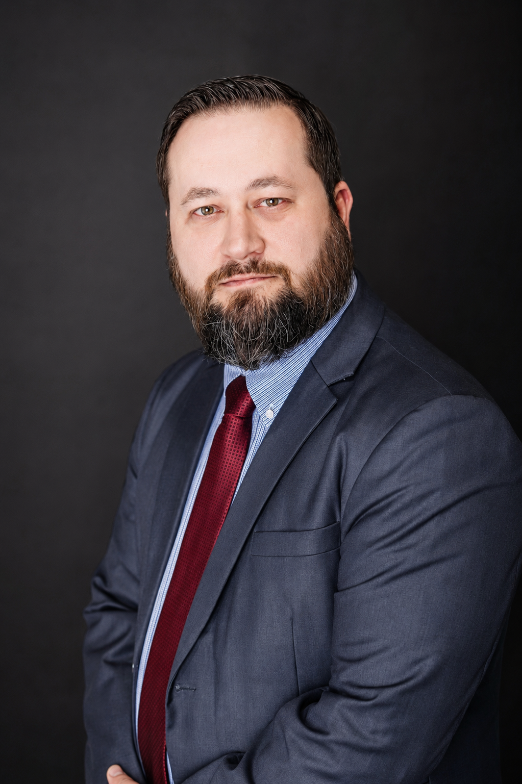A professional headshot of a man with a beard and mustache, wearing a dark suit jacket, light blue shirt, and red tie, posing against a black background.
